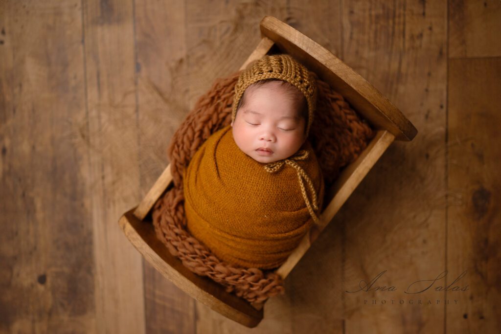 newborn baby wrapped in a brown cheesecloth and bonnet laying in a wooden bed during their Newborn Photo Session in Edmonton