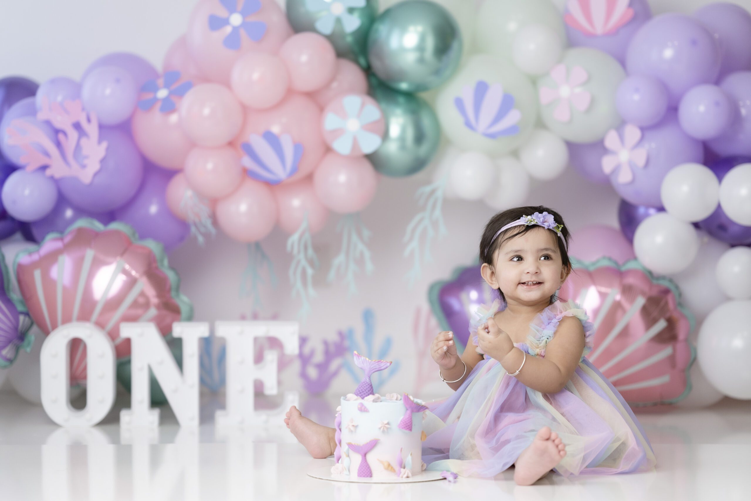 A little girl in a purple and yellow dress sits on the floor with a mermaid themed cake for her cake smash photoshoot in Edmonton