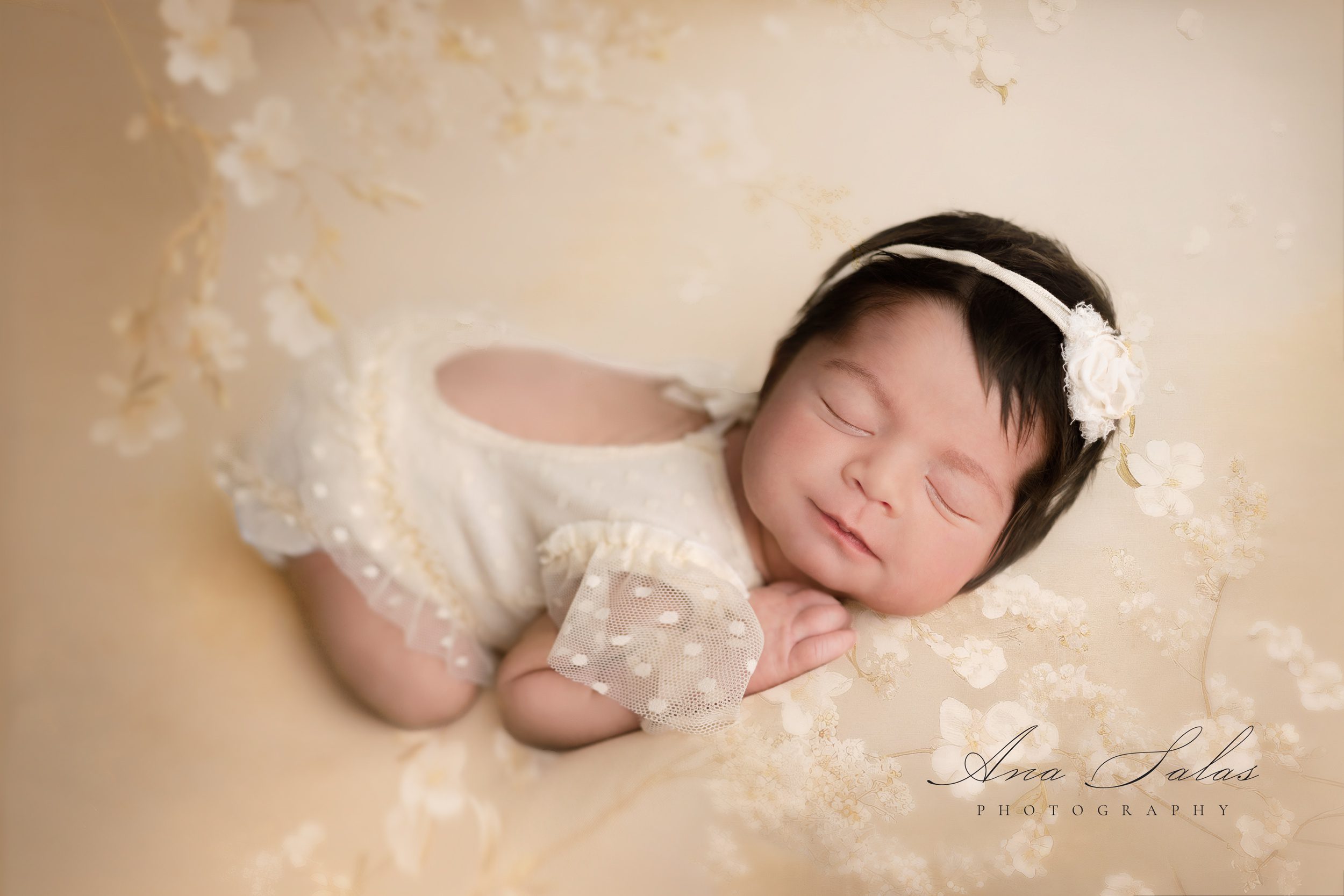 A newborn baby girl happily sleeps in a white dress in a studio