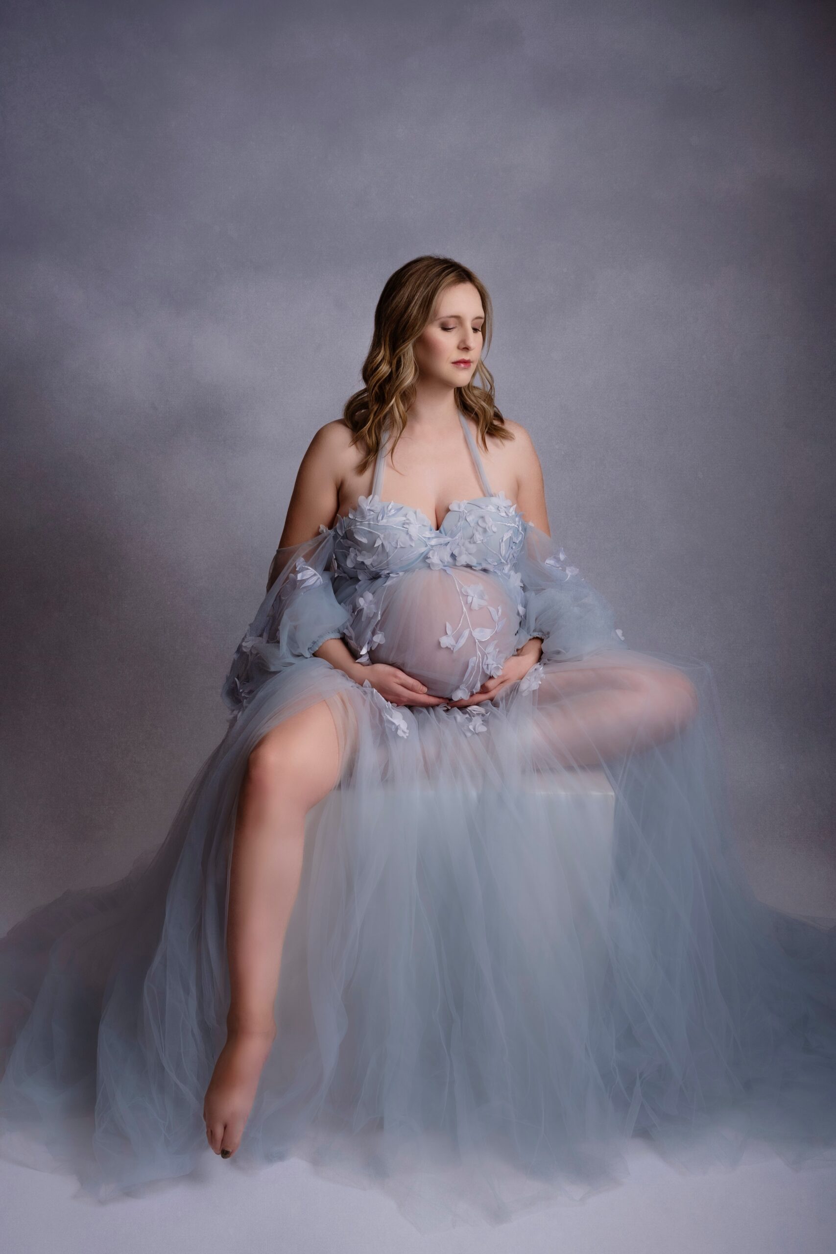 A mom to be in a blue maternity gown holds her bump while sitting on a box in a studio before meeting edmonton midwives