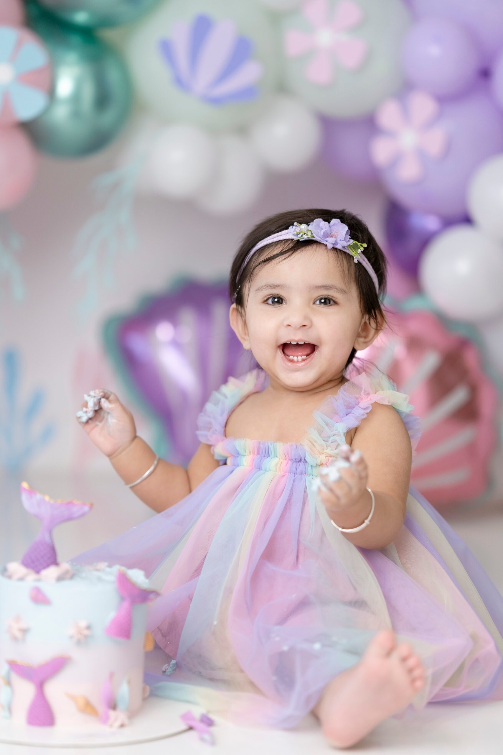 A happy toddler girl in a colorful dress happily smashes her mermaid themed cake before some Mommy and Me classes Edmonton