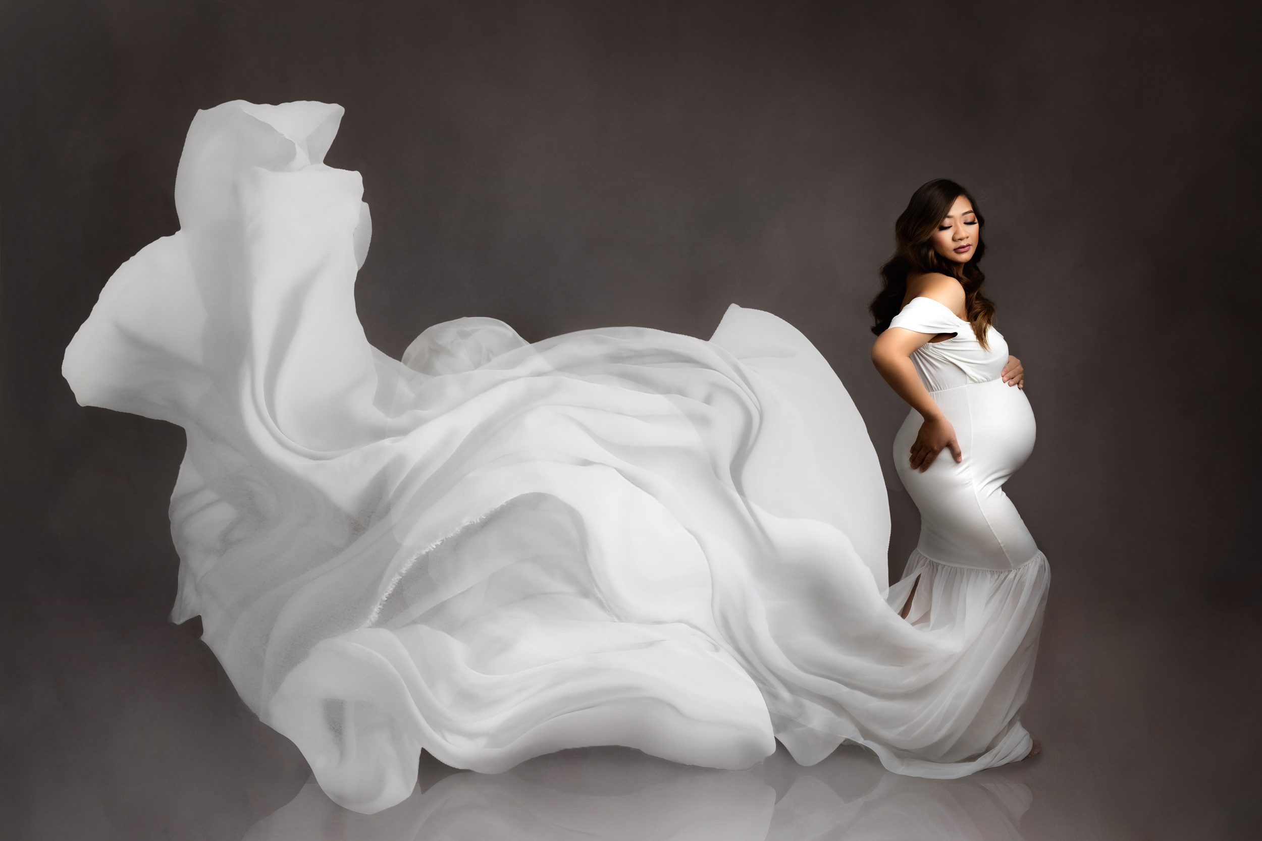 A mom to be stands in a studio holding her bump as the train of her maternity gown flows behind her