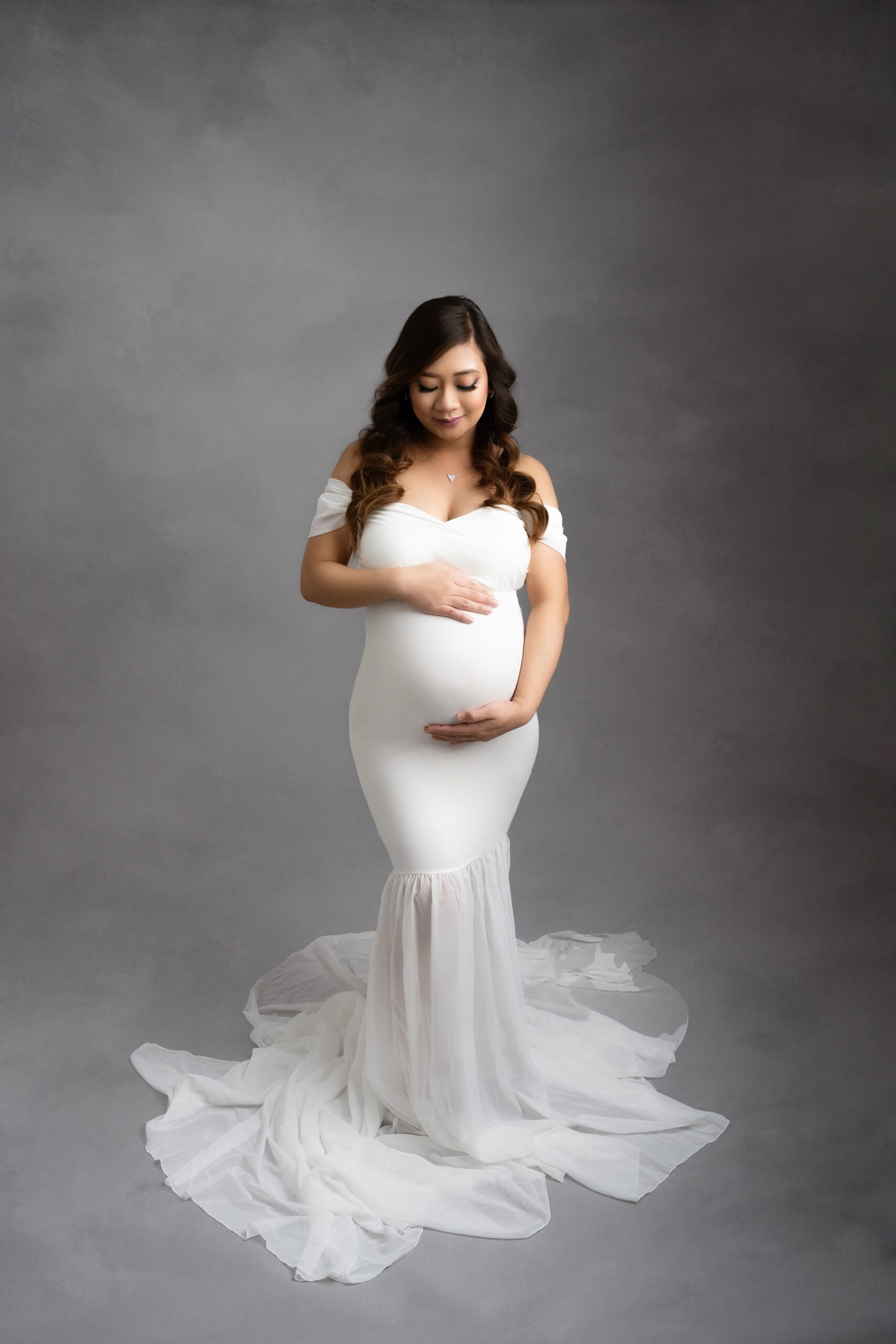 A mother to be in a white maternity gown smiles down to her hands on the bump while standing in a studio before placenta encapsulation edmonton