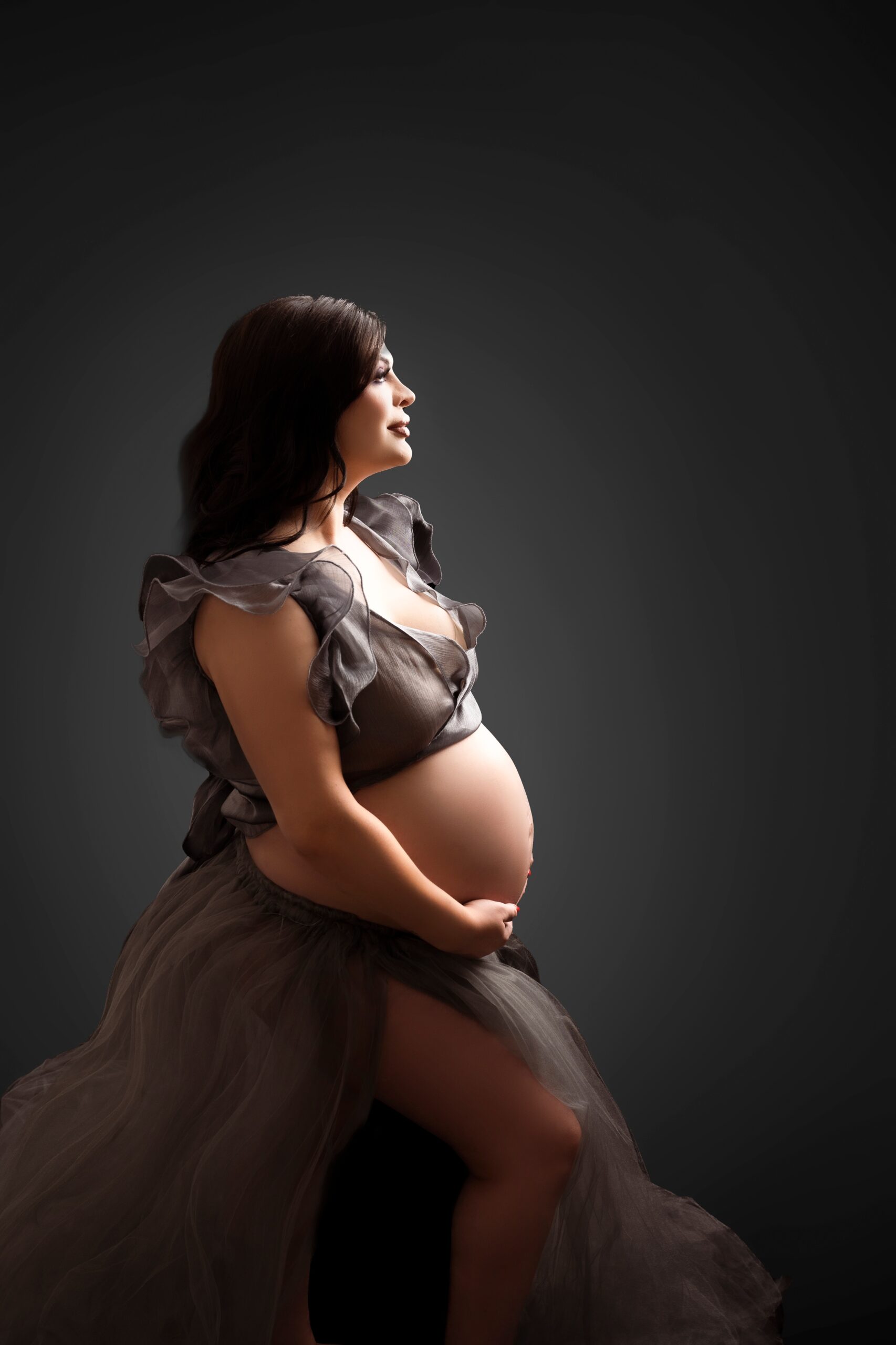 A mother to be stands in a light in a studio holding her exposed bump while wearing a grey maternity gown after visiting baby shower venues edmonton