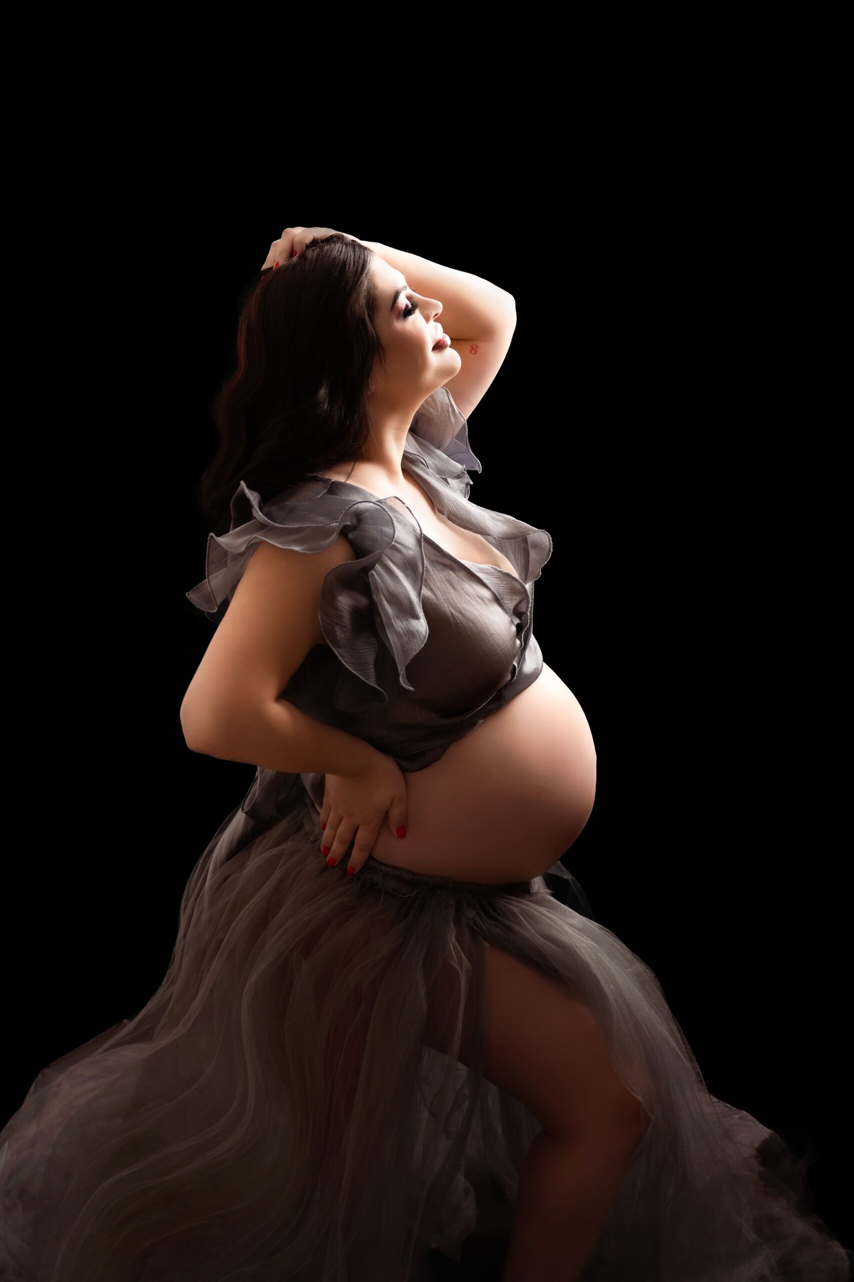 A mom to be stands proudly in a studio in a grey maternity gown with the bump out after looking at baby shower venues edmonton