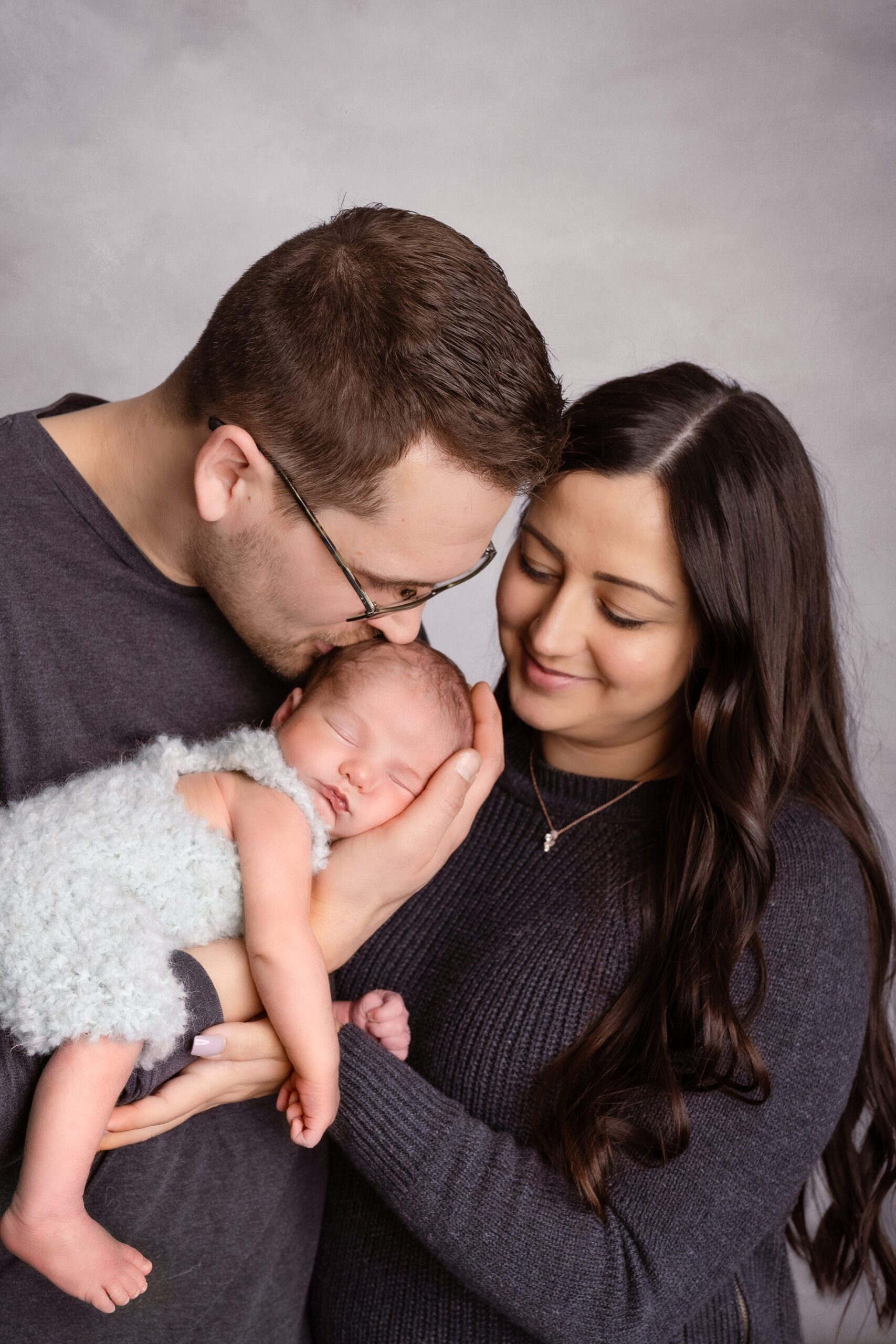 A father cradles and kisses his sleeping newborn baby with mom leaning in