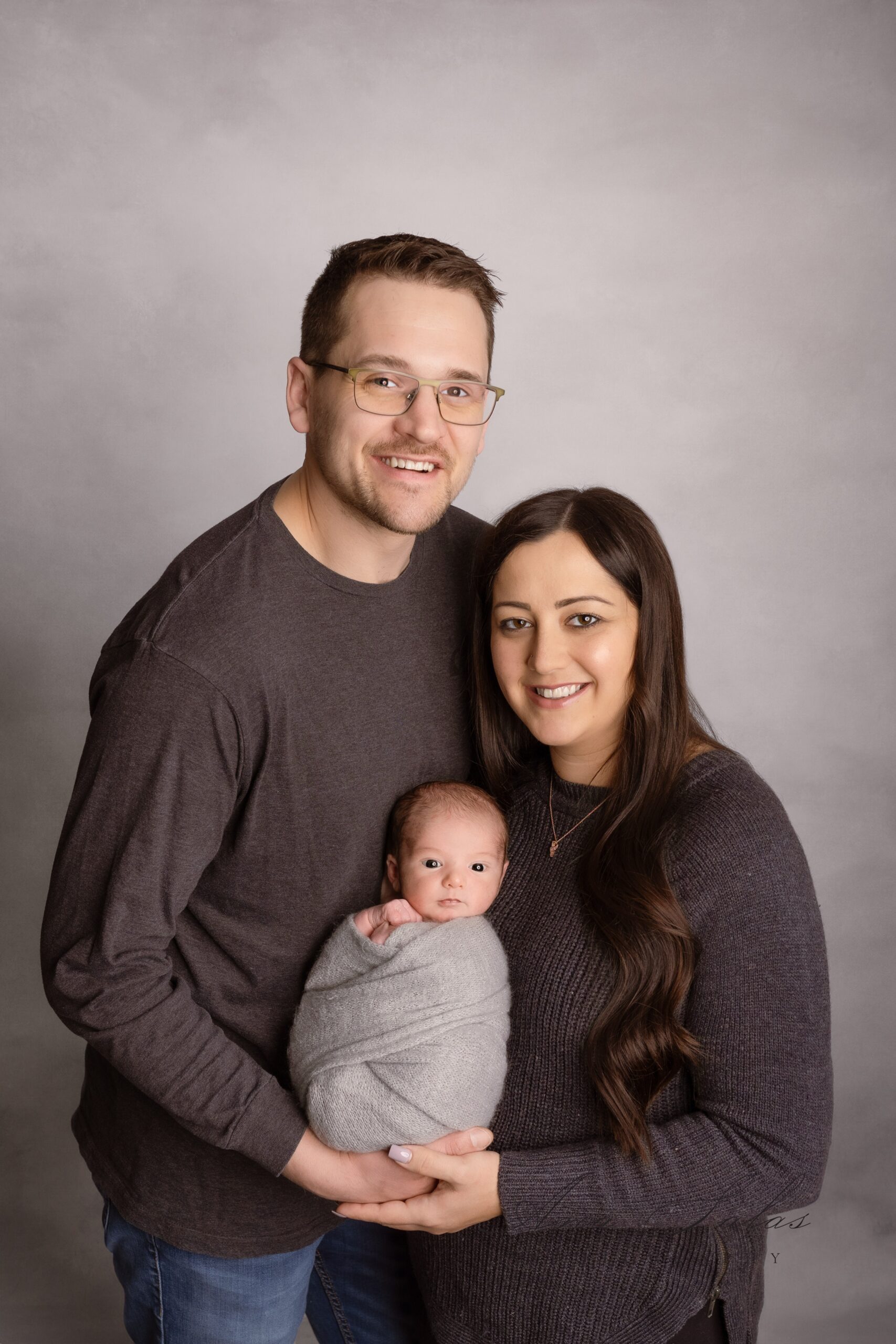 Happy new parents stand in matching sweaters while holding their newborn baby in a grey swaddle in a studio after some edmonton parenting classes