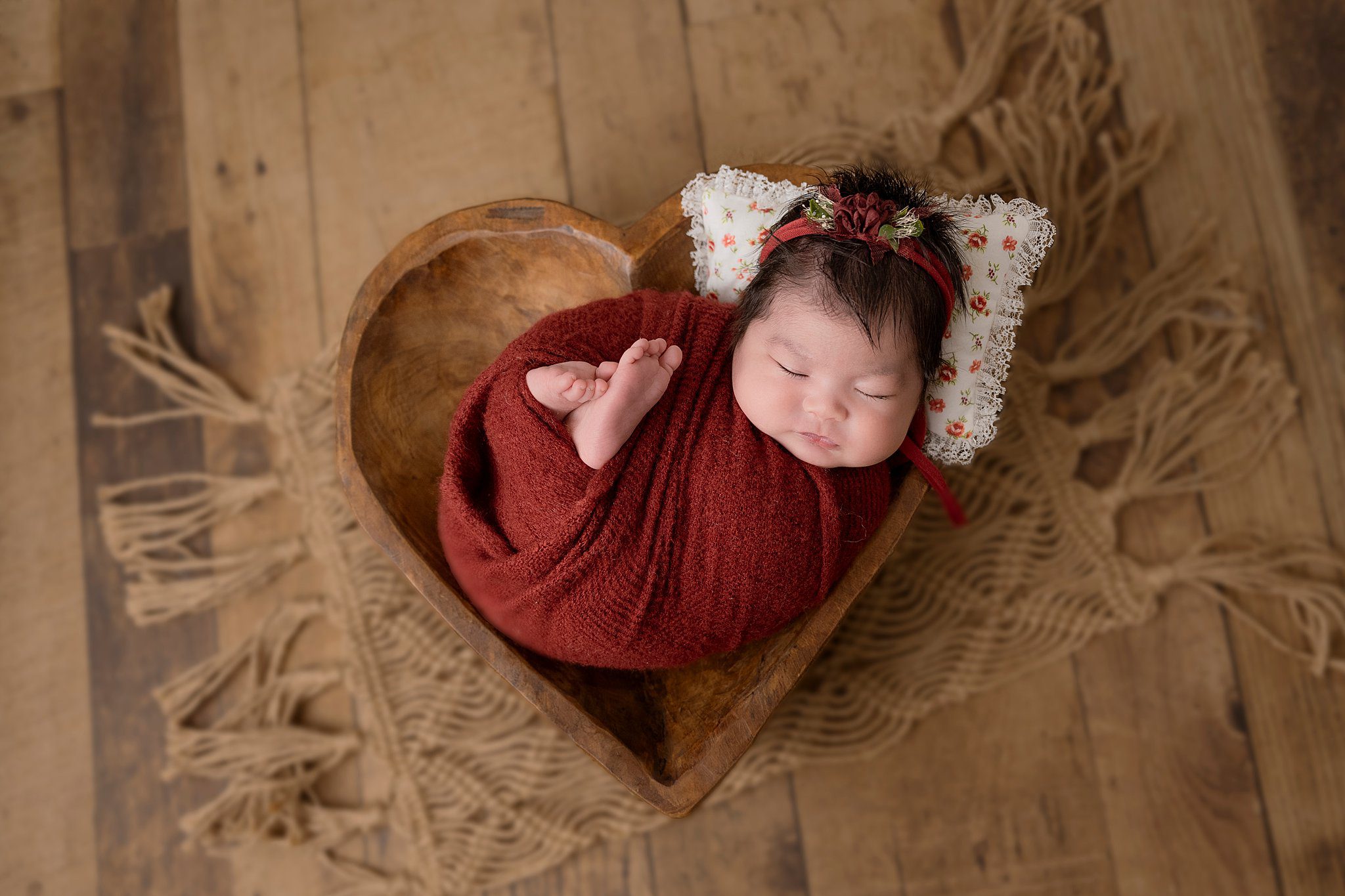 A newborn baby girl sleeps in a red knit swaddle in a wooden bowl shaped like a heart