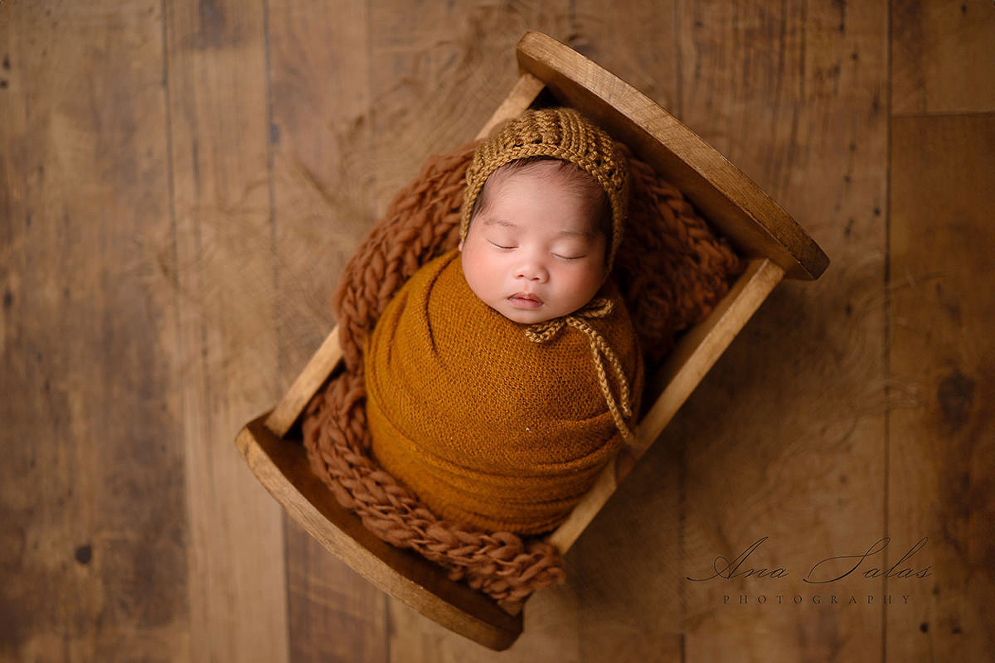 A newborn baby sleeps in a brown knit swaddle and bonnet in a wooden crib in a studio before visiting edmonton pediatric dentists