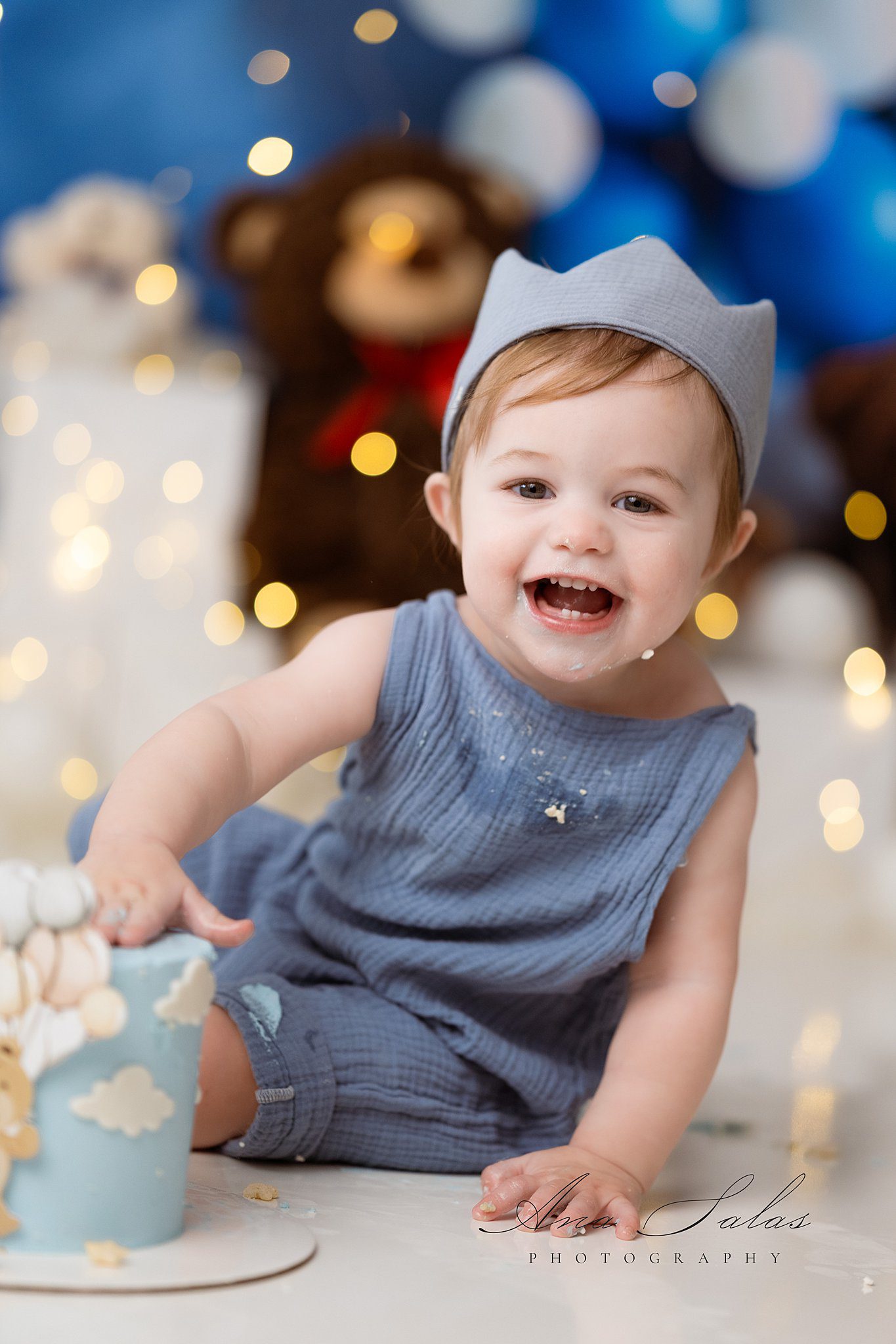 A happy toddler boy smashes his birthday cake in a blue onesie covered in cake