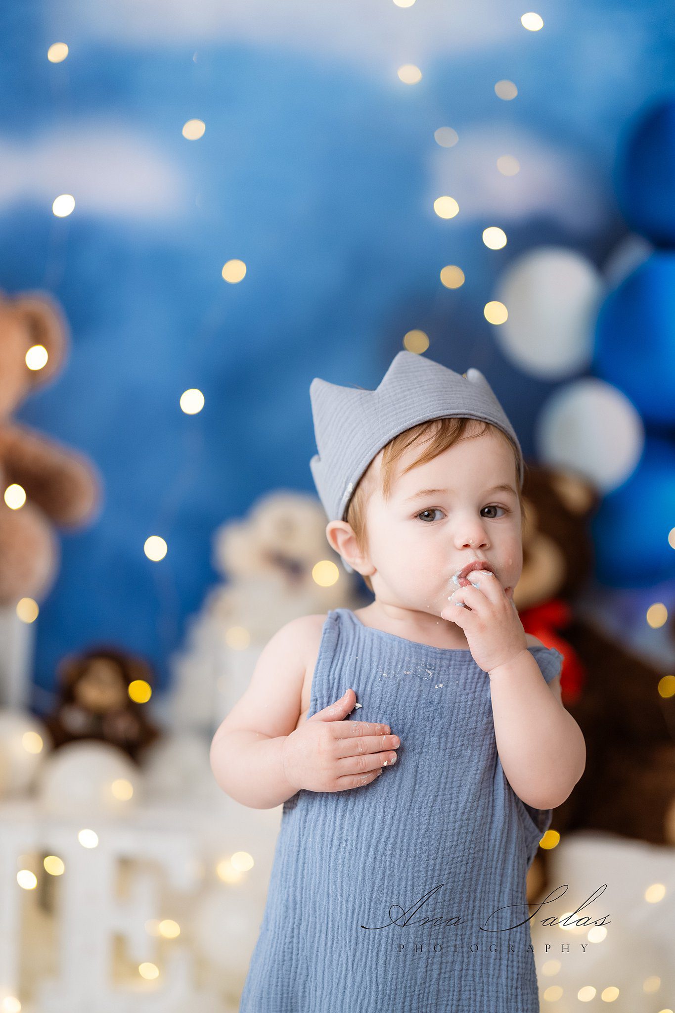 A toddler boy in a blue onesie and crown eats cake off his fingers while standing in a studio after visiting edmonton pediatricians