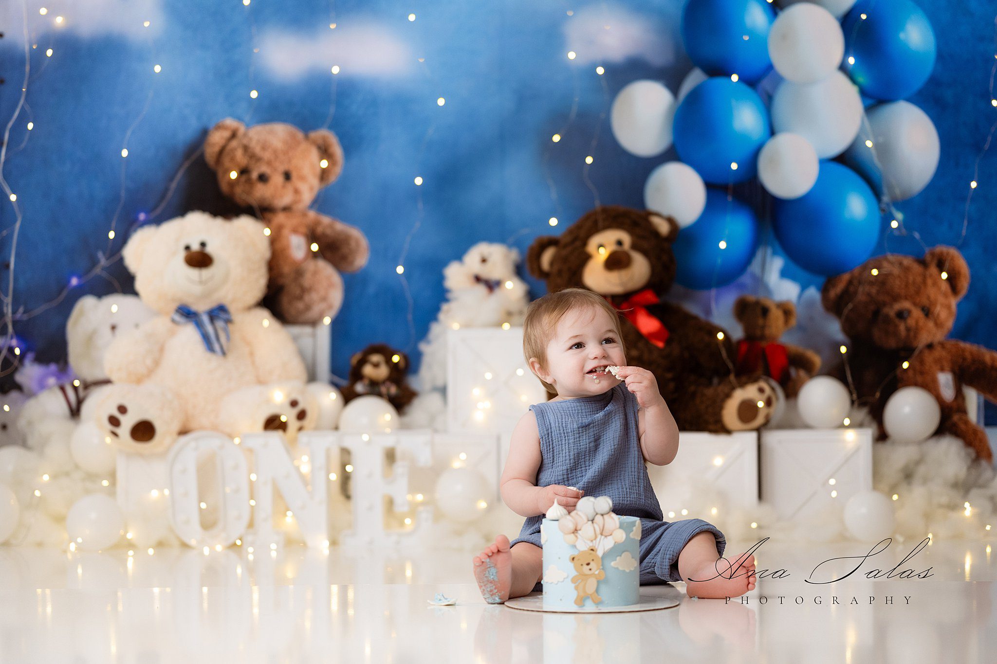 A happy toddler boy in a blue onesie sits with his birthday cake in a bear themed studio after visiting edmonton pediatricians