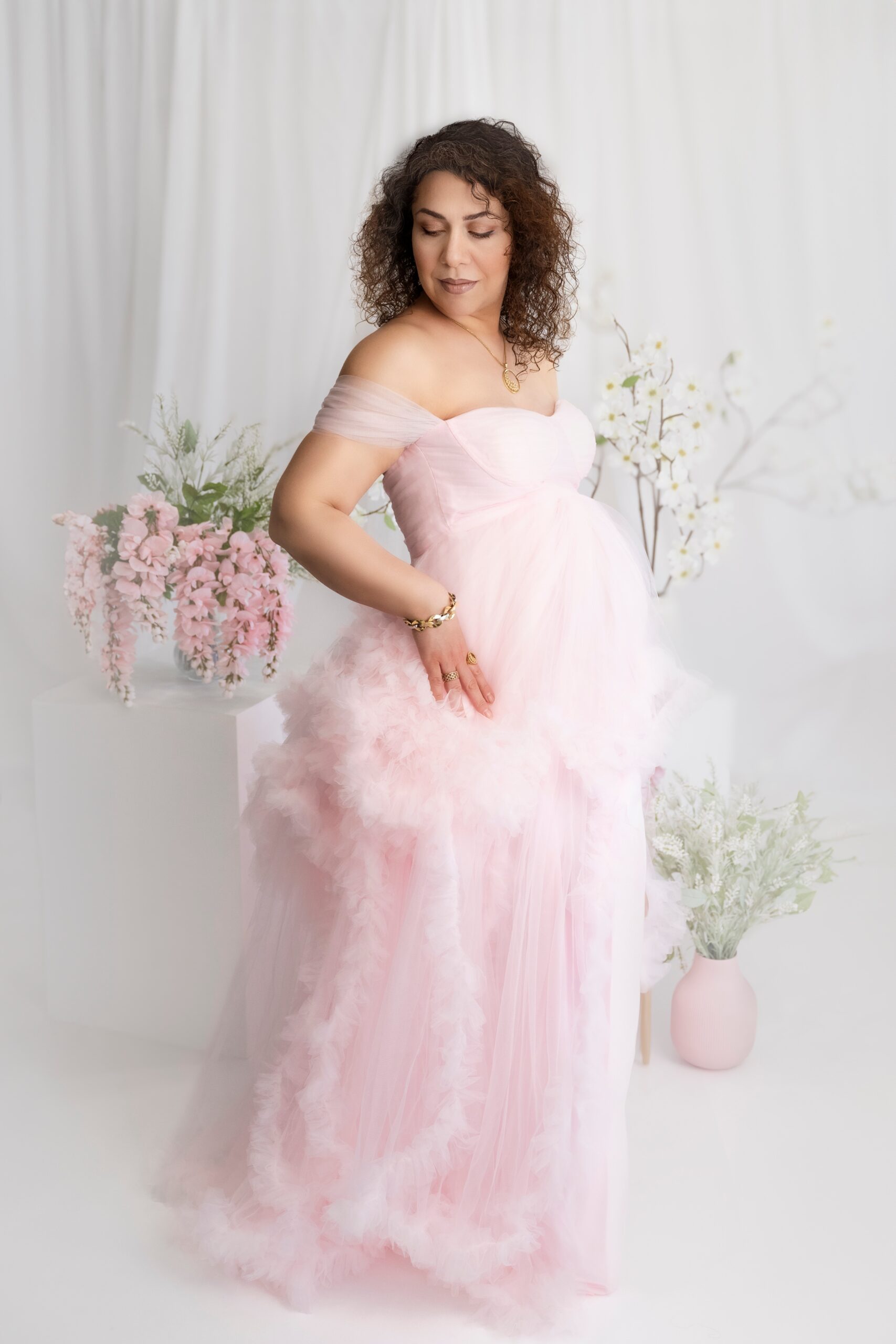 A mom to be in a pink maternity gown stands in a studio gazing down her shoulder after some pregnancy acupuncture edmonton