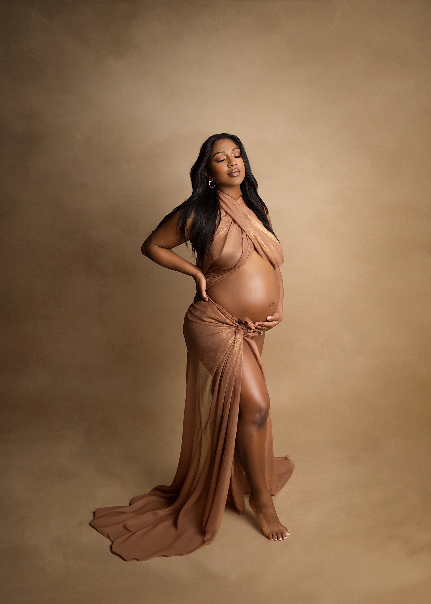 A mother to be stands in a studio in a brown wrap holding her bump