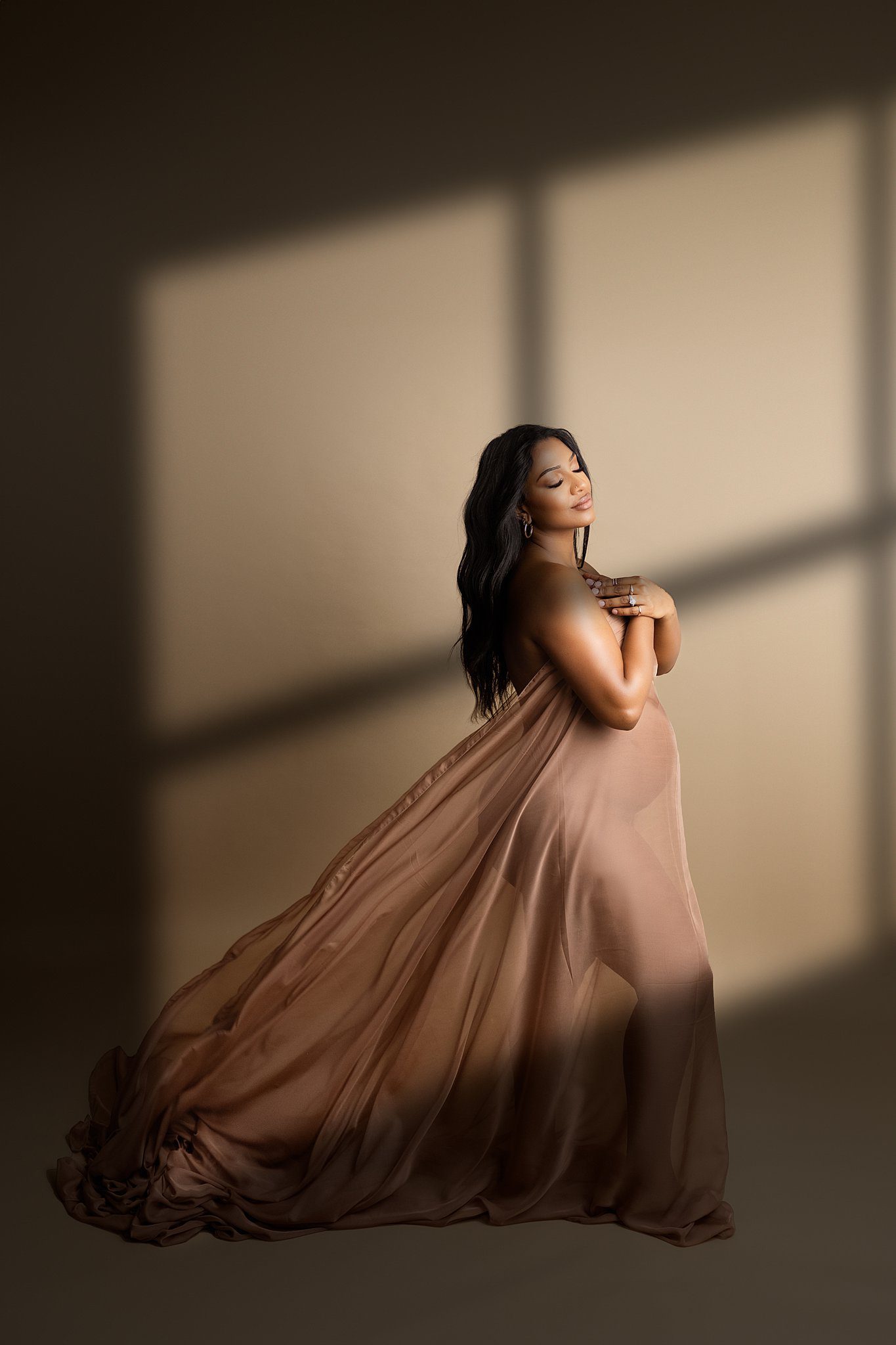 A mother to be stands in the light of a window covering with a sheer brown fabric in a studio before visiting a prenatal chiropractor edmonton