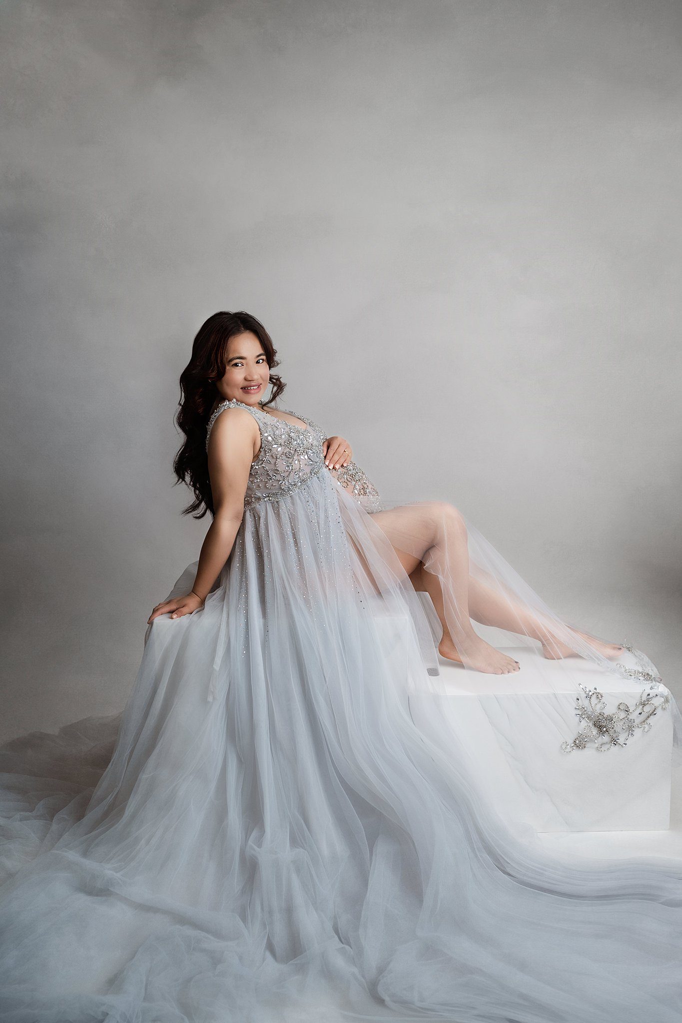 A happy mother to be sits on white boxes in a studio wearing a long flowing blue maternity gown