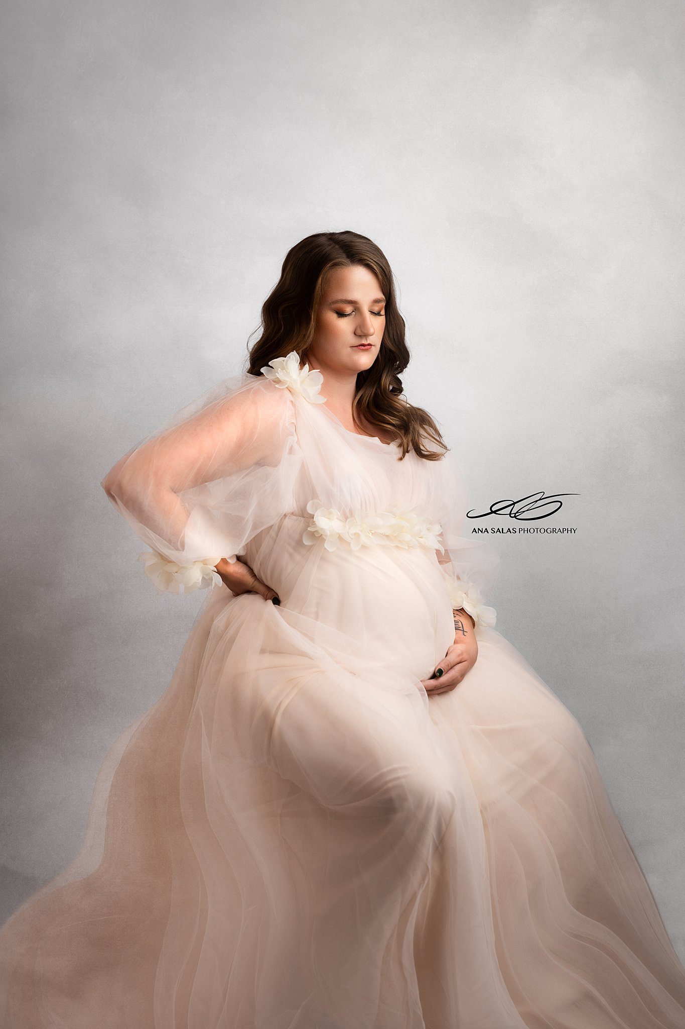 A mother to be in a long flowing white maternity gown gazes down at her bump while sitting in a studio before a prenatal massage edmonton