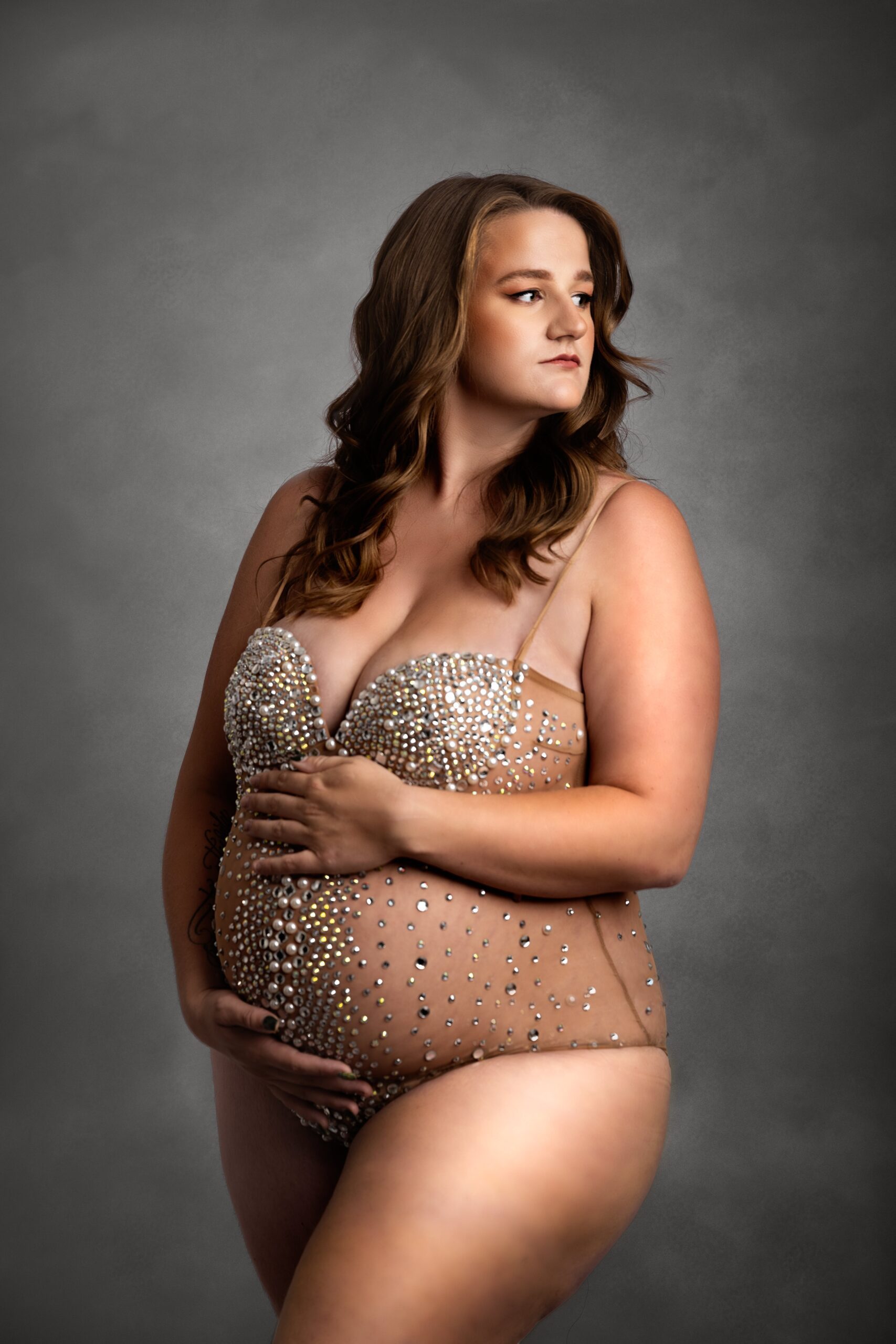 A mom to be in an ornate bodysuit stands in a studio gazing over her shoulder and holding the bump after some prenatal yoga edmonton