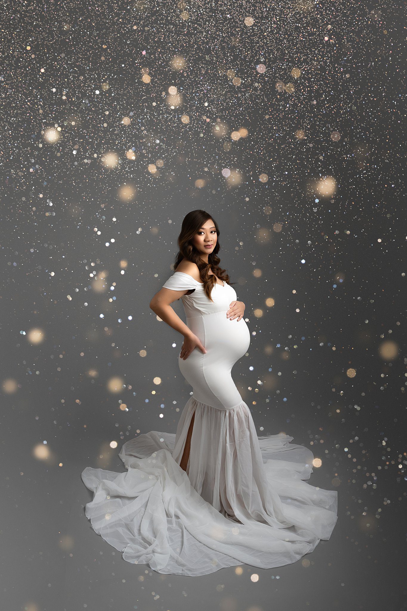 A mom to be stands under a shower of glitter in a white maternity gown with a hand on top of her bump after her 3d ultrasound edmonton