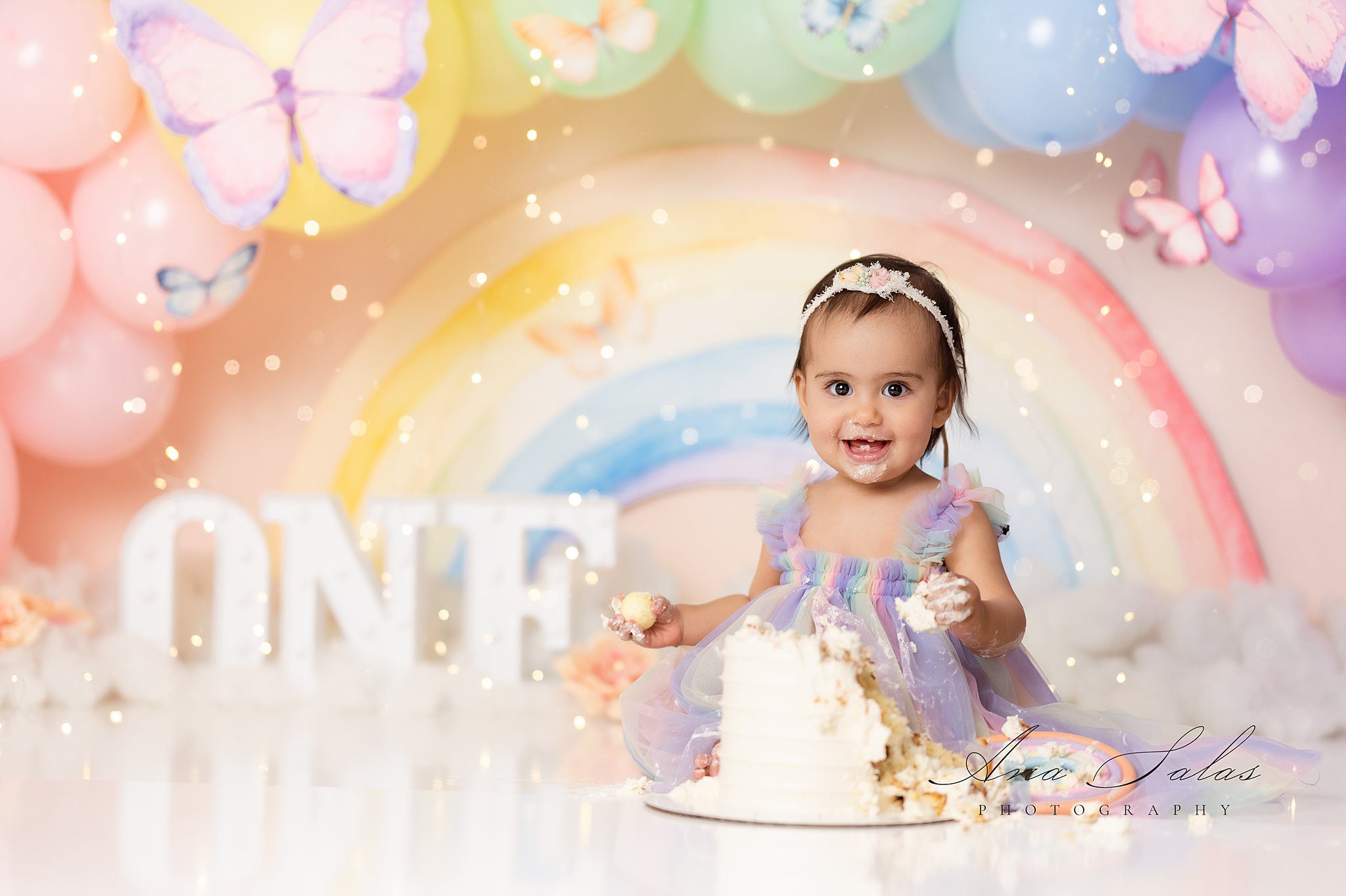 A smiling toddler in a rainbow dress celebrates her first birthday with a cake smash