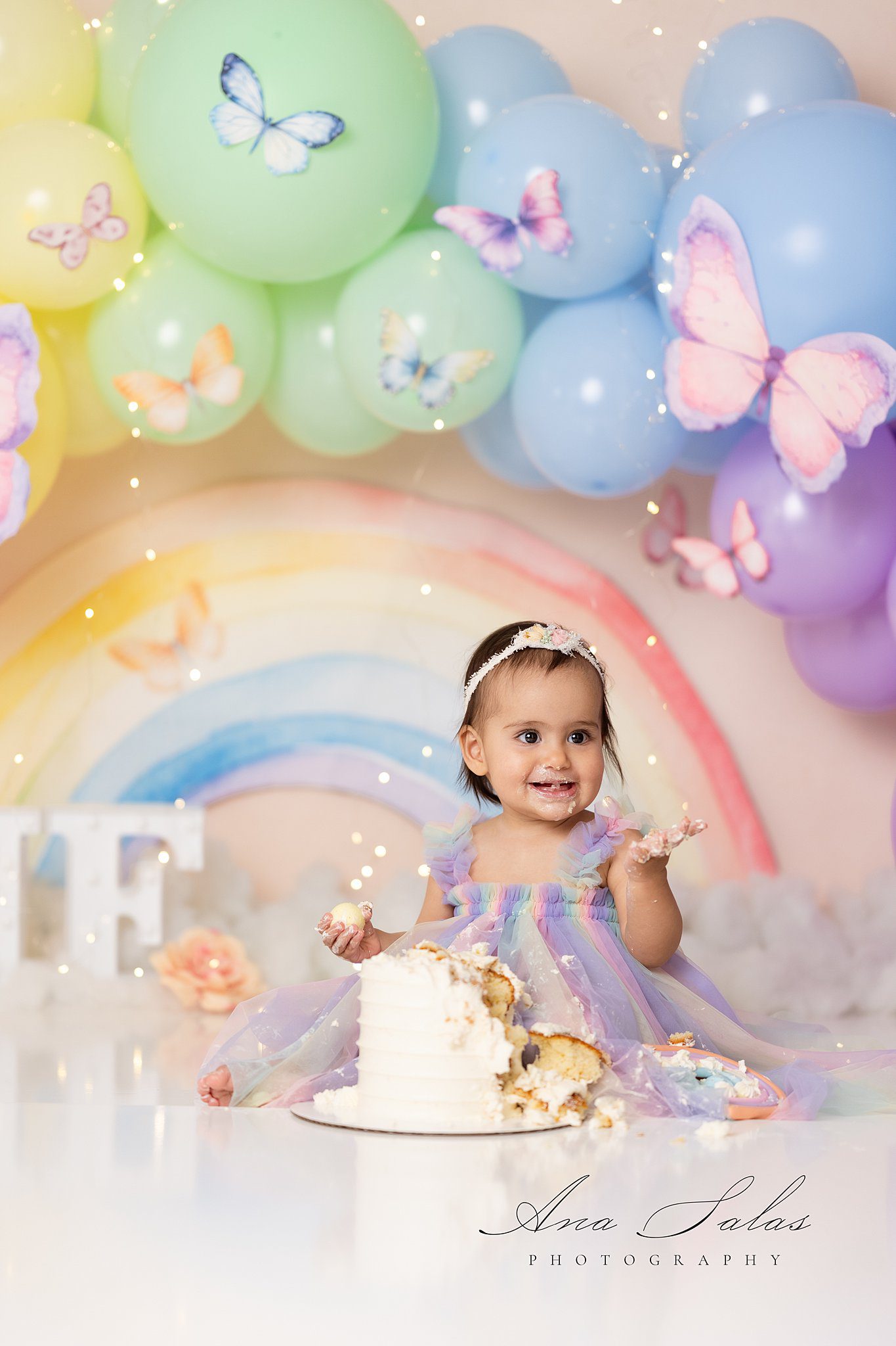 A toddler girl in a rainbow dress happily smashes and eats her birthday cake in a studio after meeting an au pair edmonton