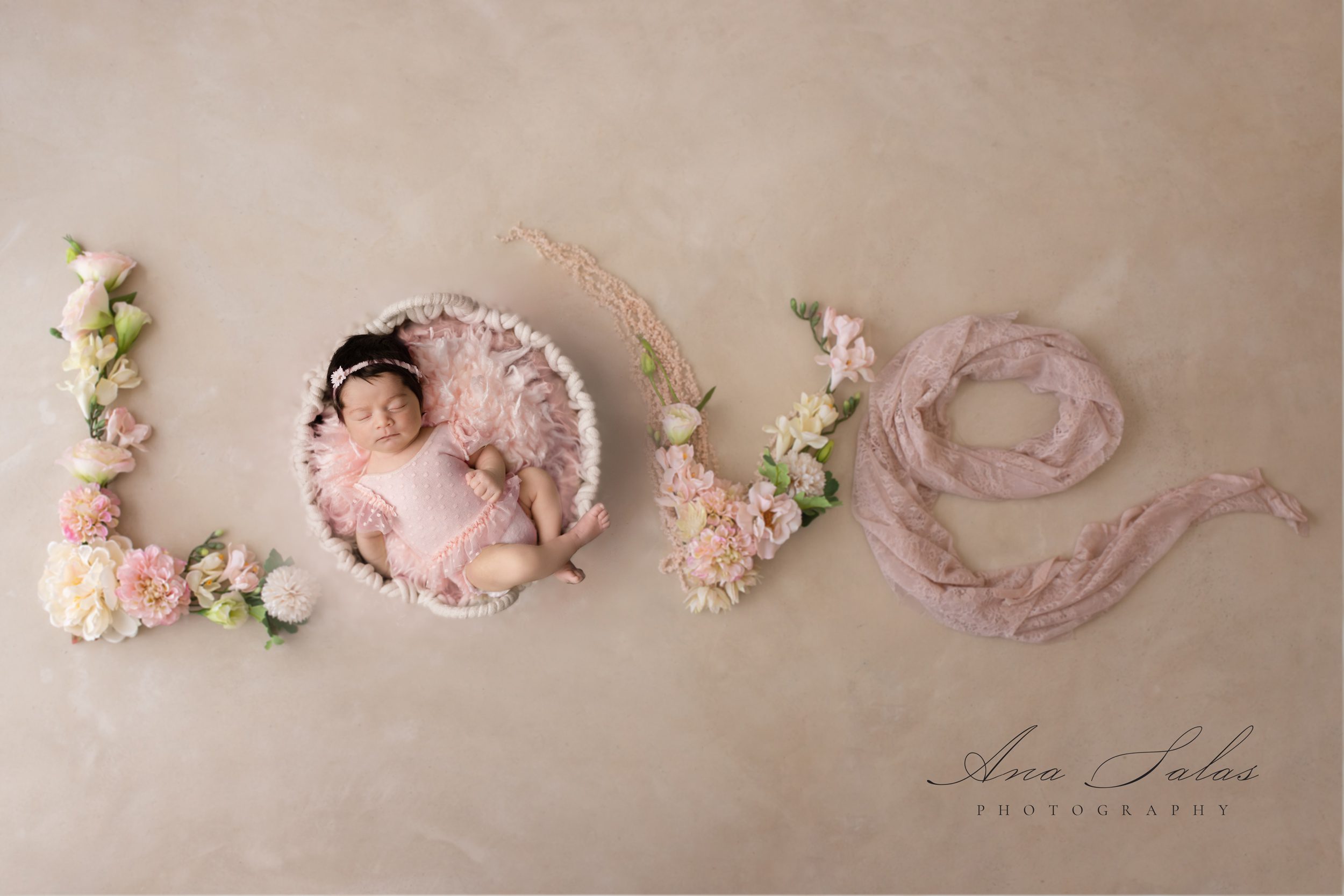 A newborn baby sleeps in a pink dress in a woven basket serving as an O for Love in flowers and fabric