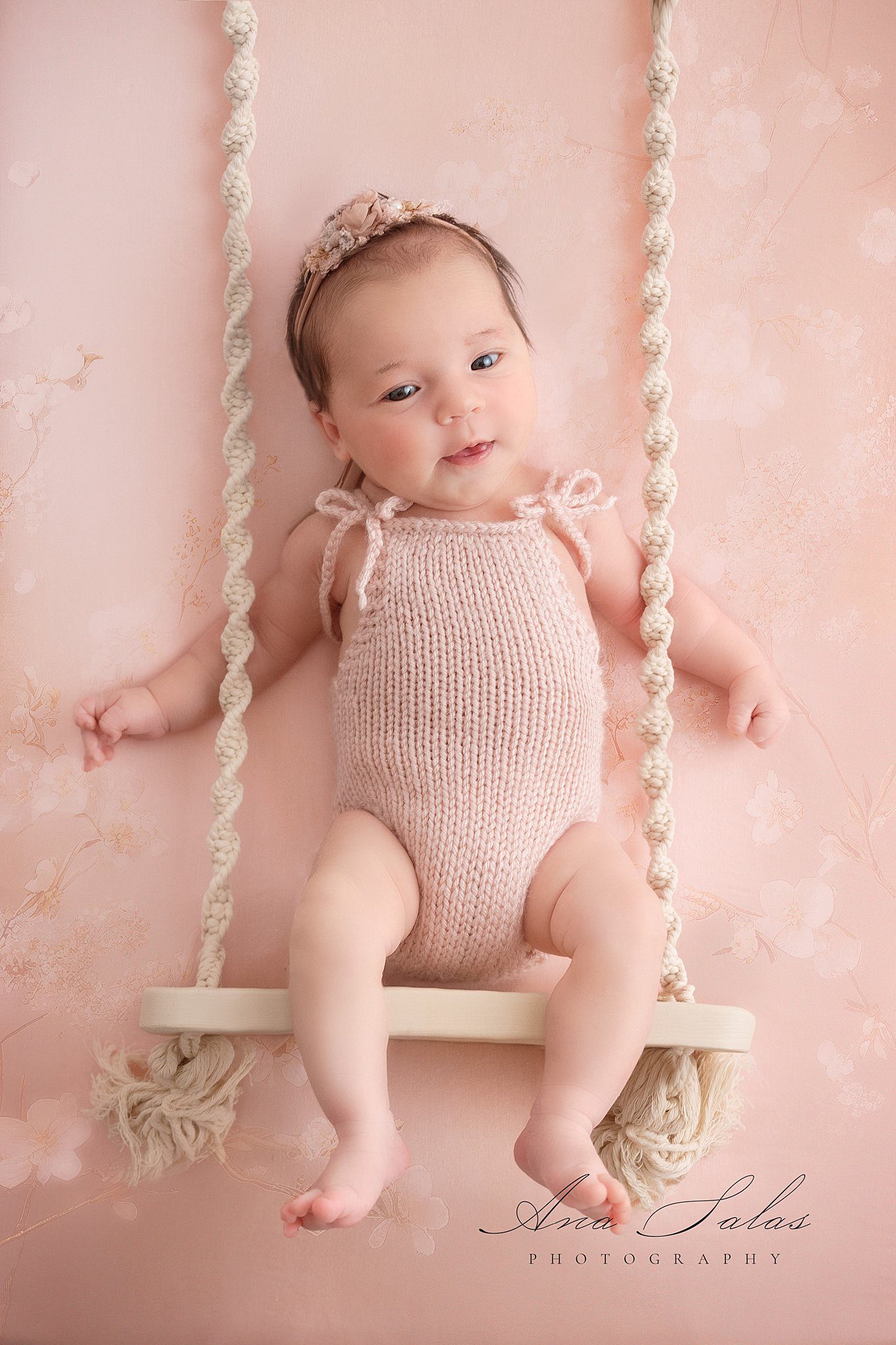 A newborn baby girl in a knit pink onesie sits on a swing in a studio with eyes open after meeting edmonton lactation consultant