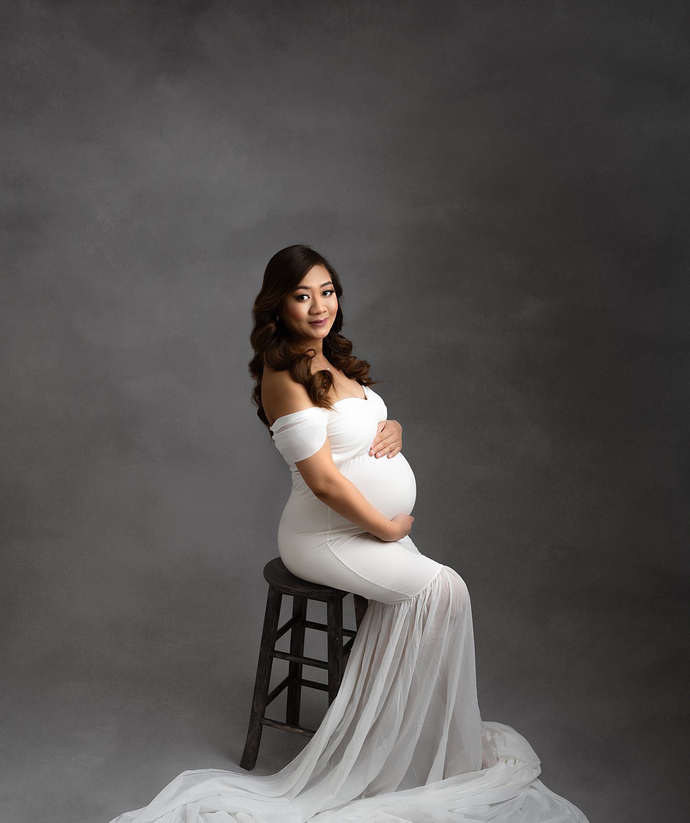 A happy mom to be sits on a wooden stool in a studio while holding her bump