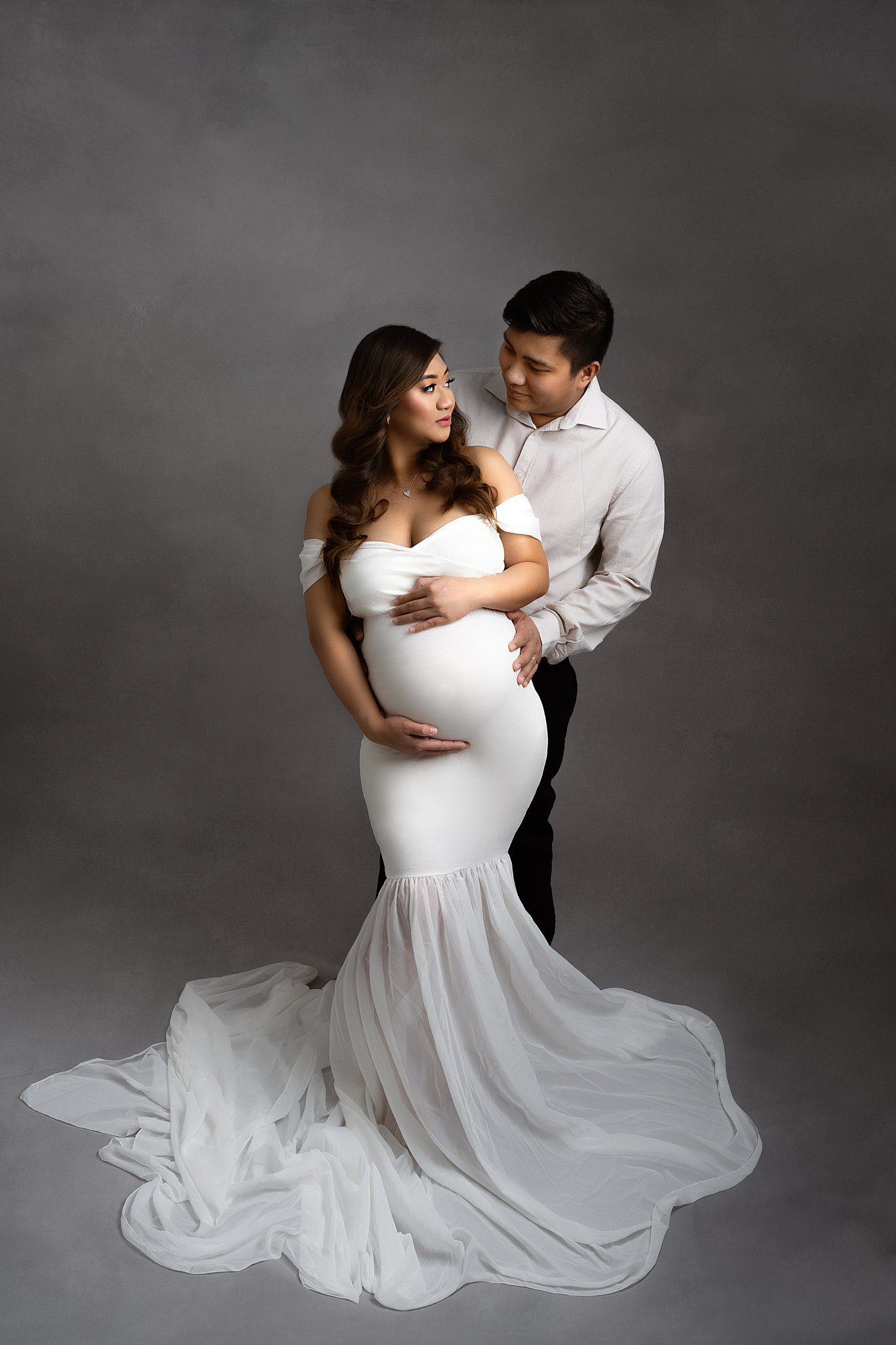 Expecting parents stand together in a studio looking at each other and holding the bump in a white maternity gown after meeting an edmonton obgyn