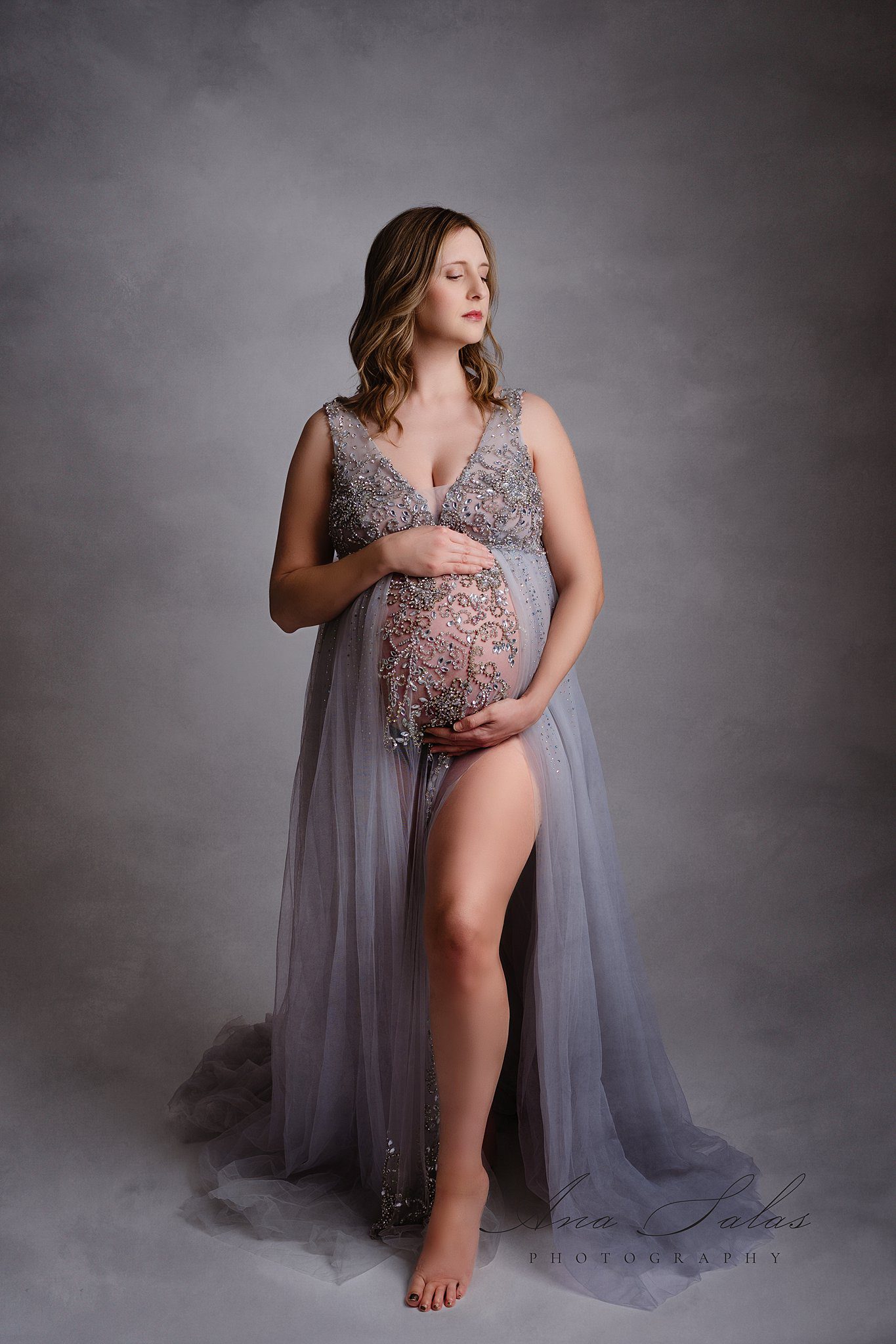 A mom to be in an ornate blue maternity gown stands in a studio with eyes closed after visiting lucina birth centre