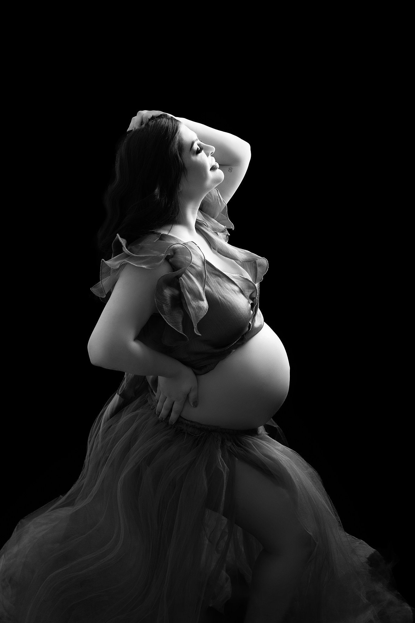 A mom to be with an arm on her hip and one in her hair stands in a studio with her bump exposed