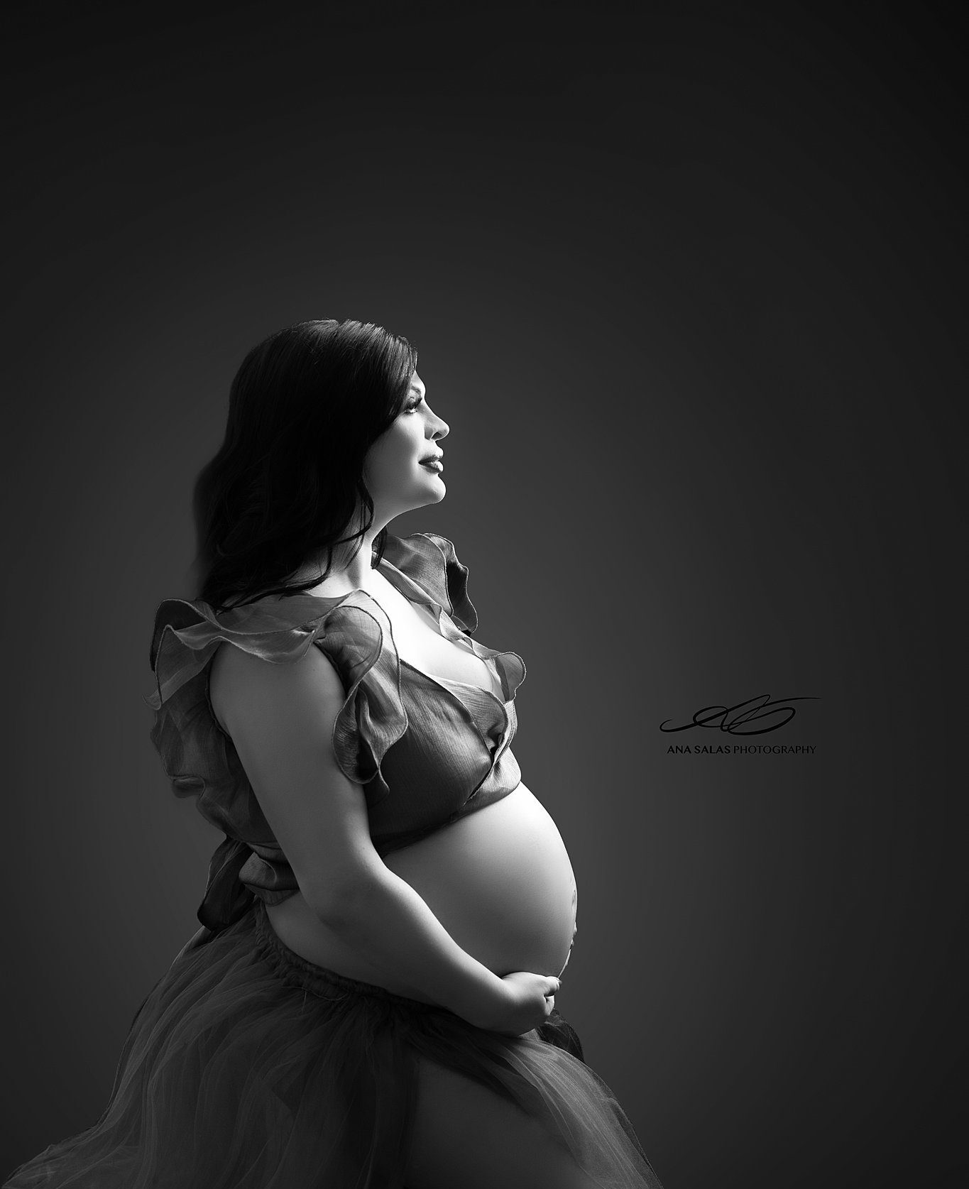 A happy mom to be sits in a spotlight in a studio in an open maternity gown after visiting lucina midwives