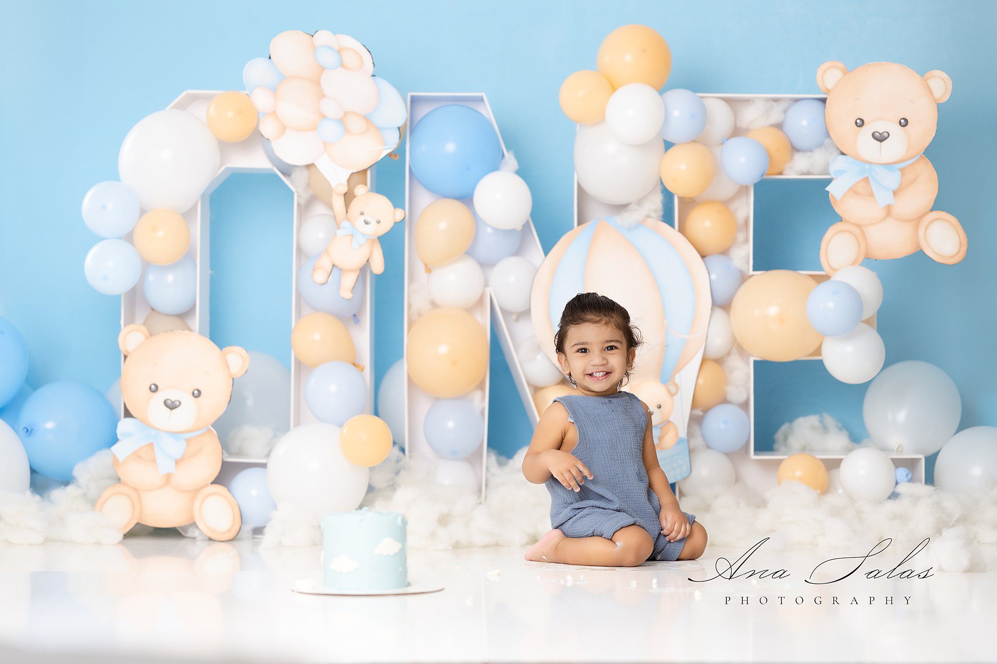 A young toddler in a blue onesie smiles while sitting with a cake