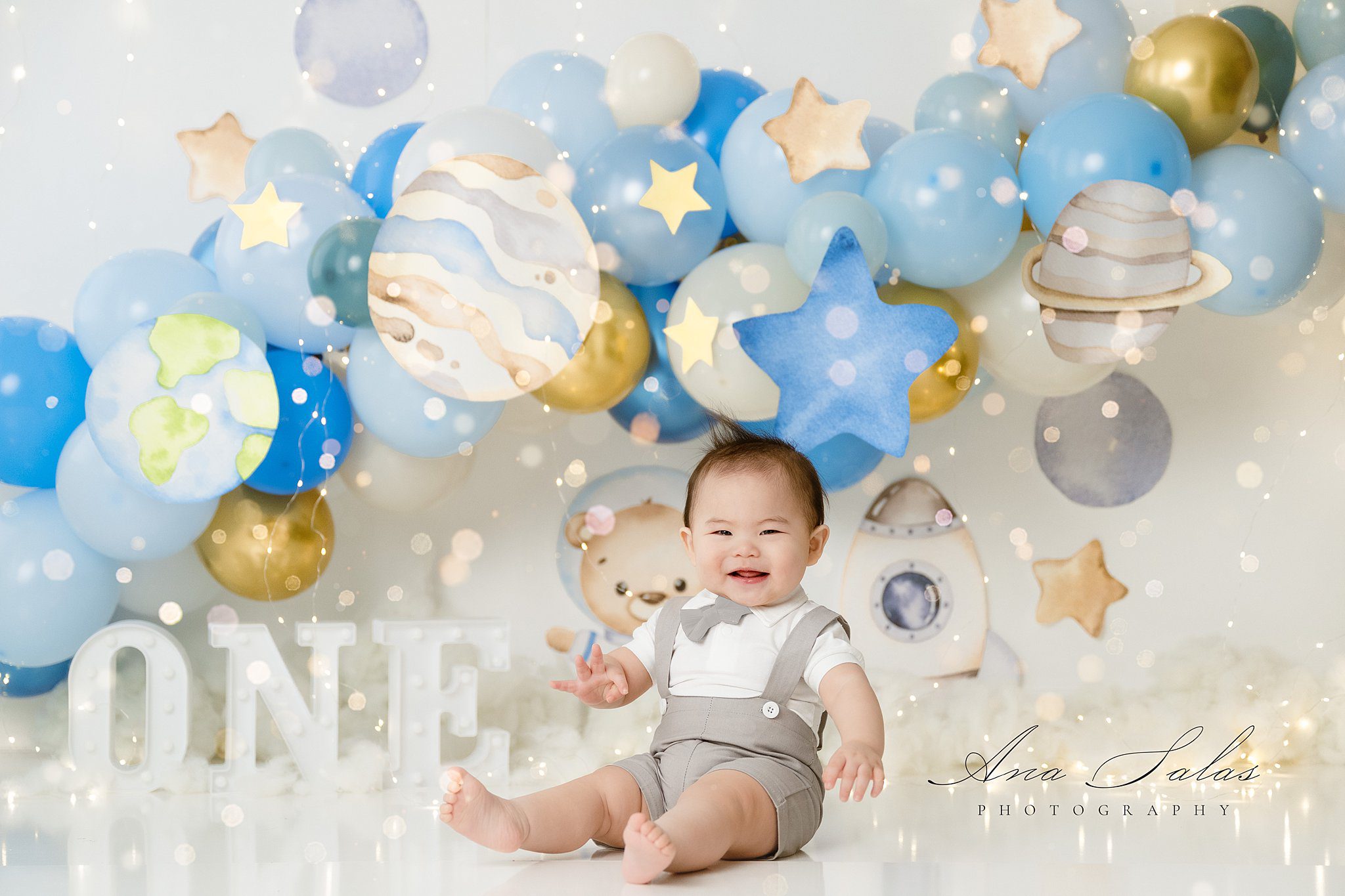 A happy toddler in overalls and matching bowtie sits in a decorated studio after meeting nannies in edmonton