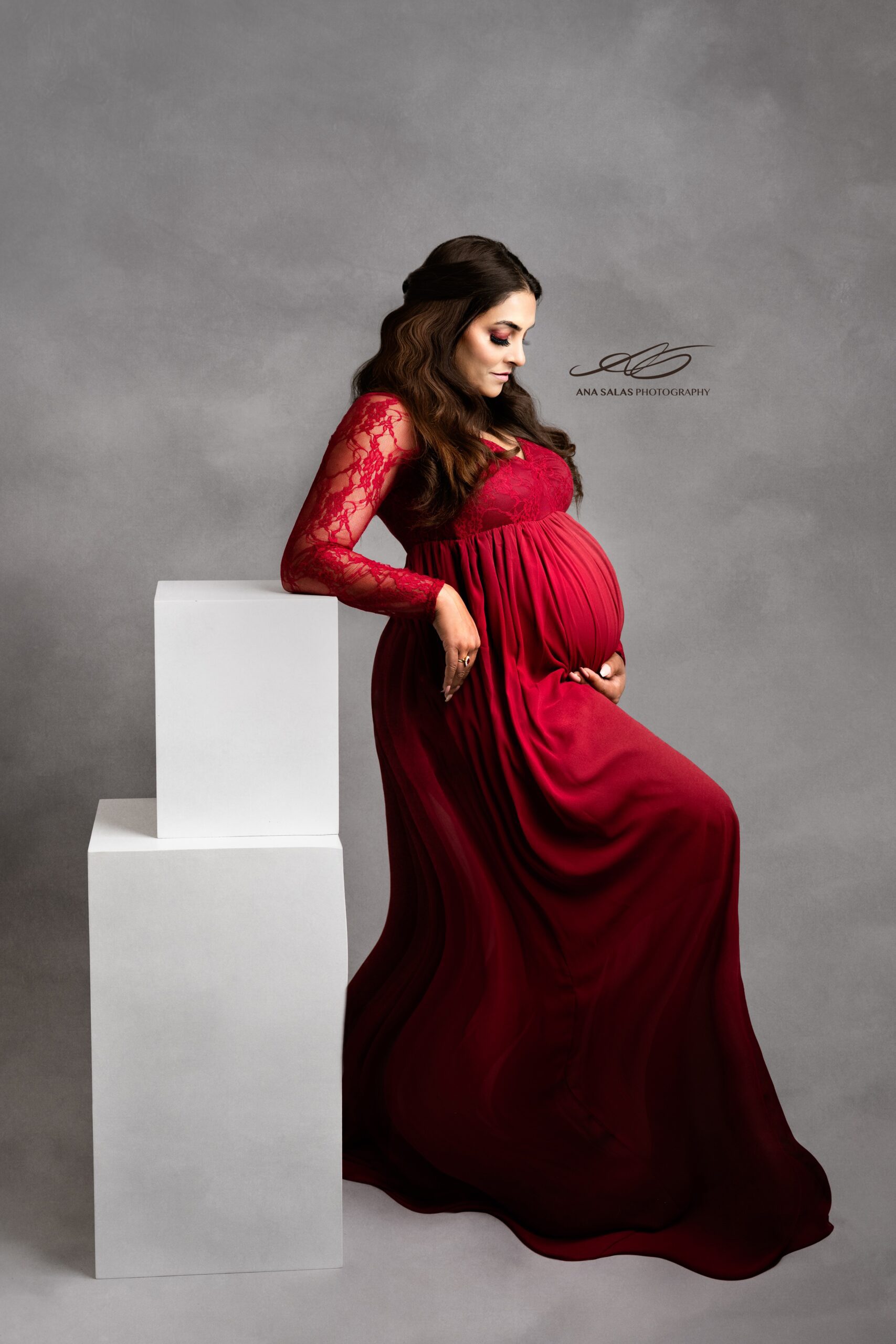 A mom to be leans on white studio boxes while smiling down at her bump in a red lace maternity gown