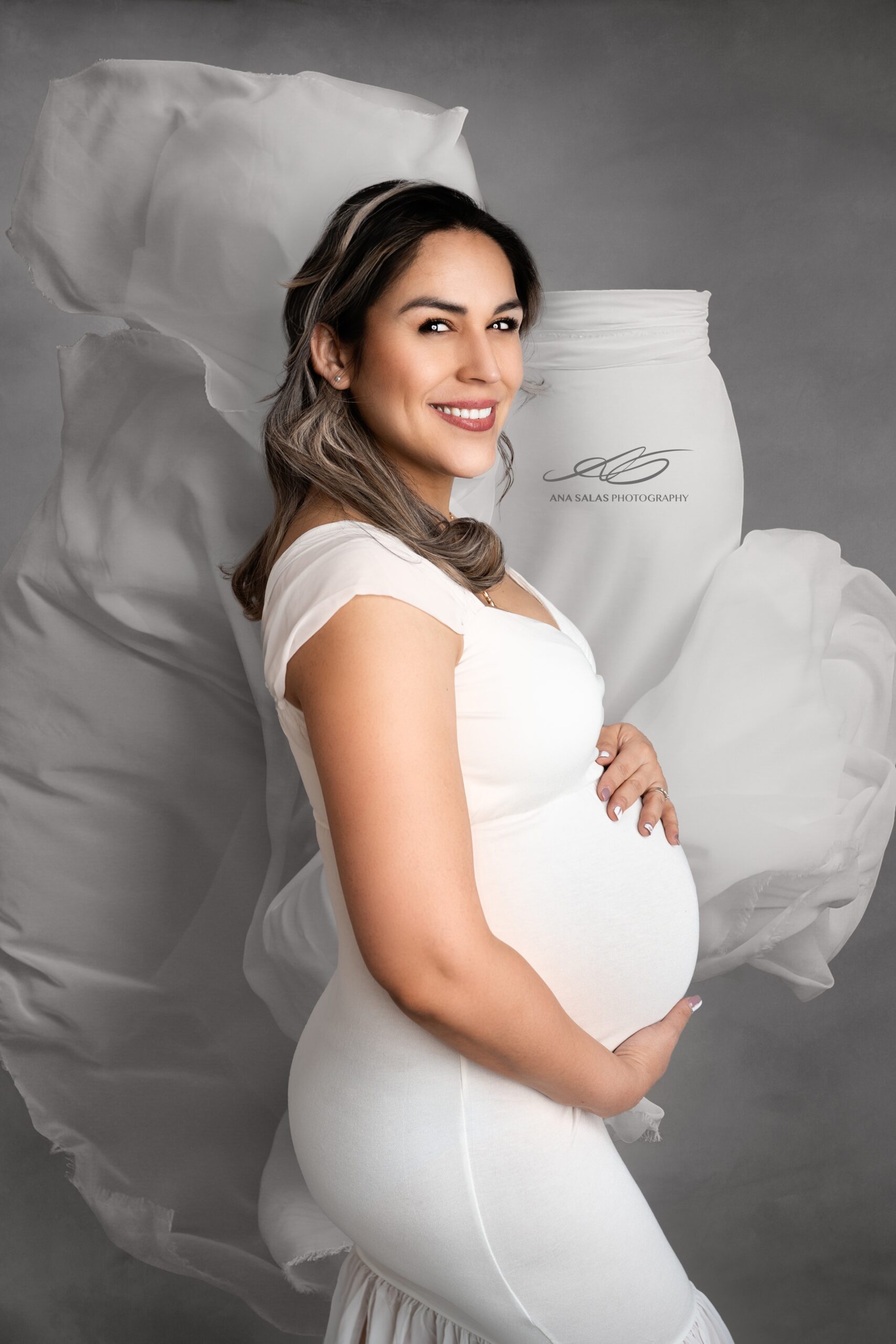 A happy pregnant woman holds her bump while smiling in a studio with the train of her white maternity gown flows behind her before her water birth in edmonton