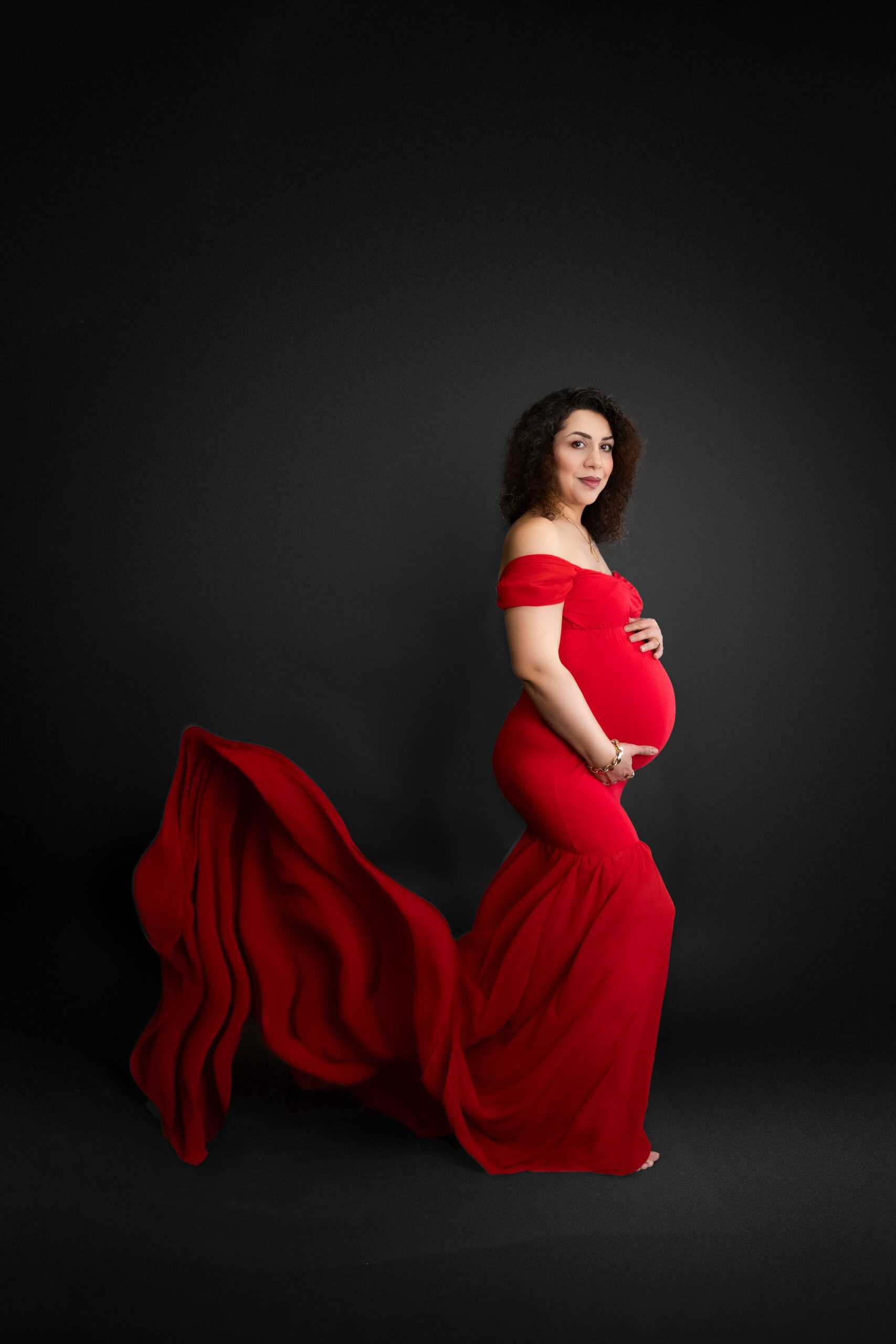 A mother to be in a red maternity gown smiles while standing in a studio with her train flowing in the wind behind her before her water birth in edmonton