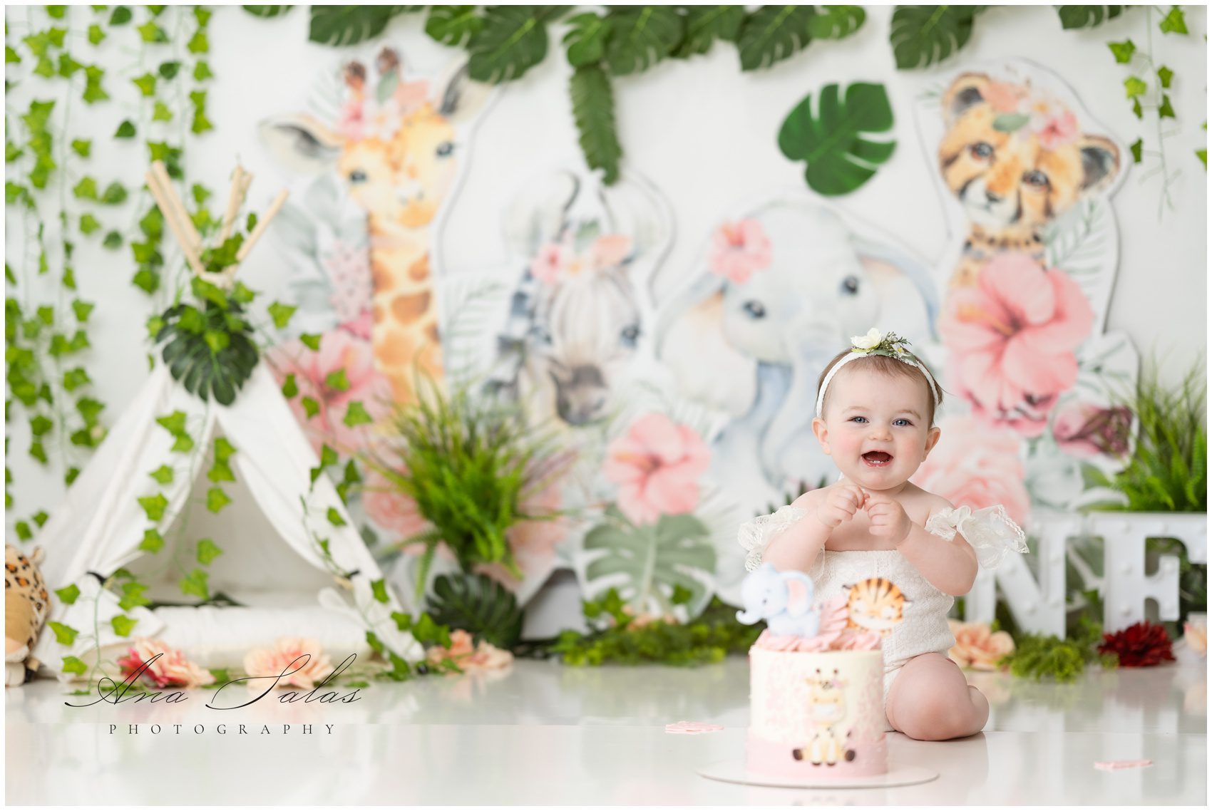 A young toddler girl in a white onesie plays with a cake in a safari themed studio