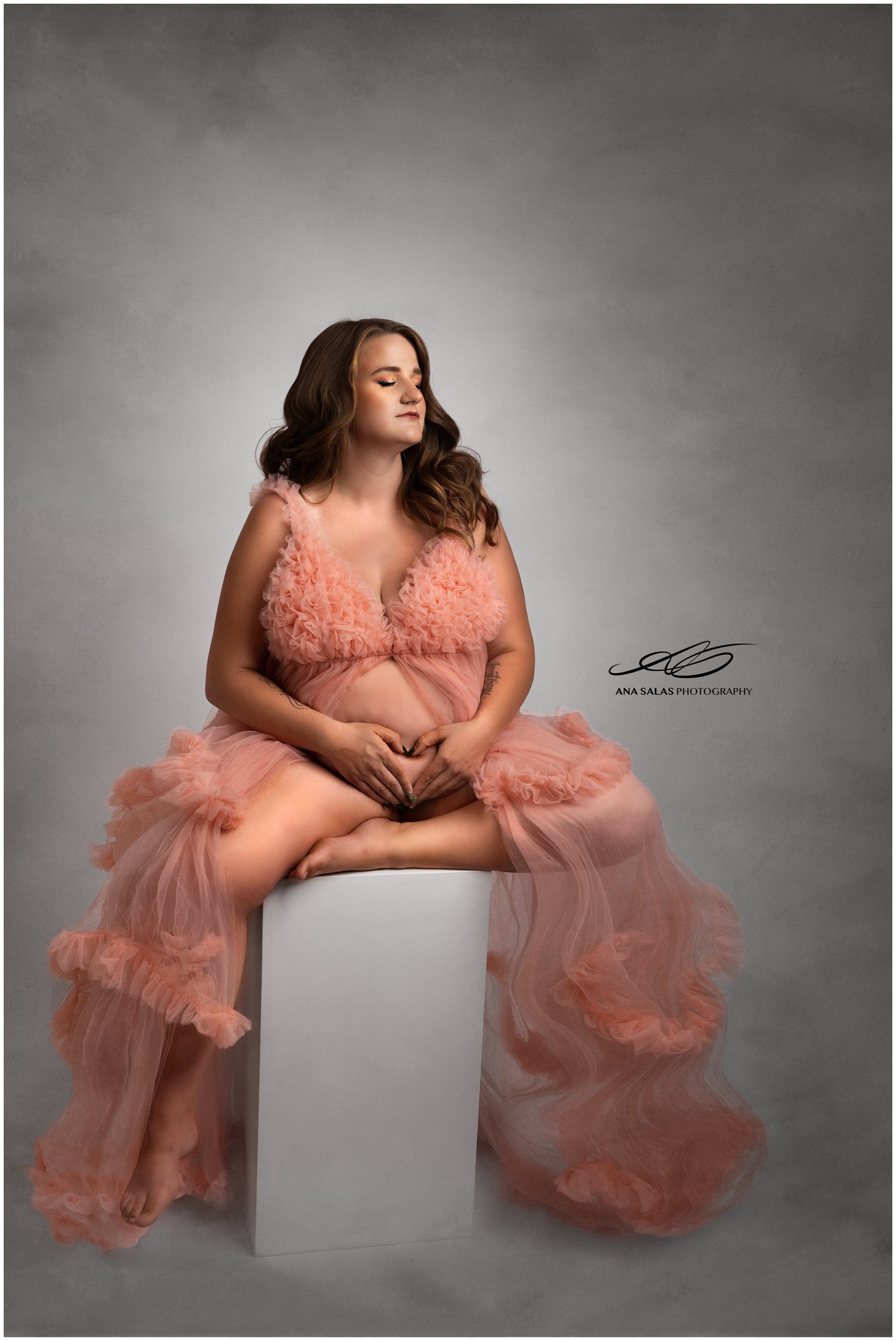 A mom to be sits on a box in a studio holding her bump in a pink maternity gown