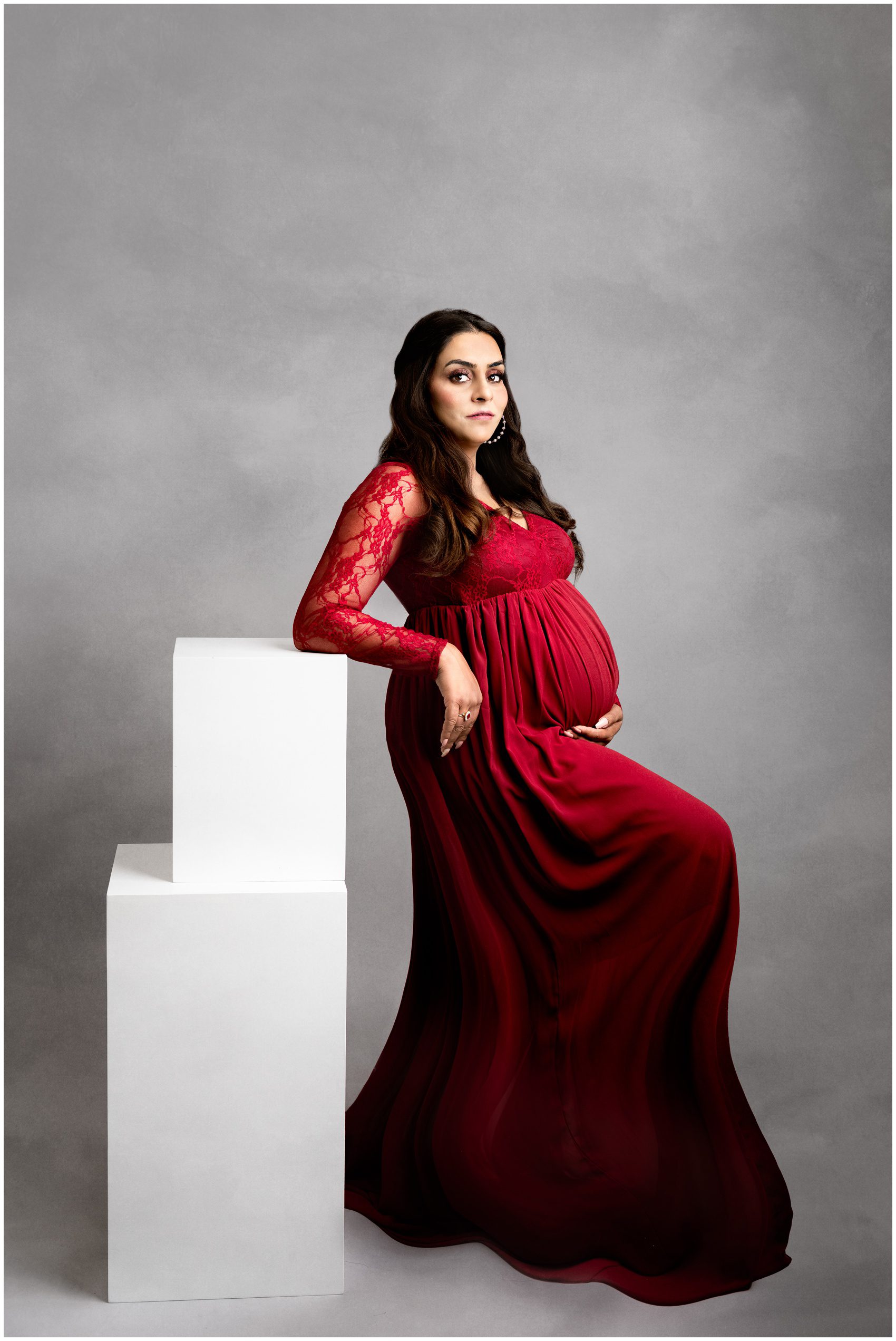 A mother to be leans on white boxes in a long red maternity gown in a studio after meeting edmonton doulas