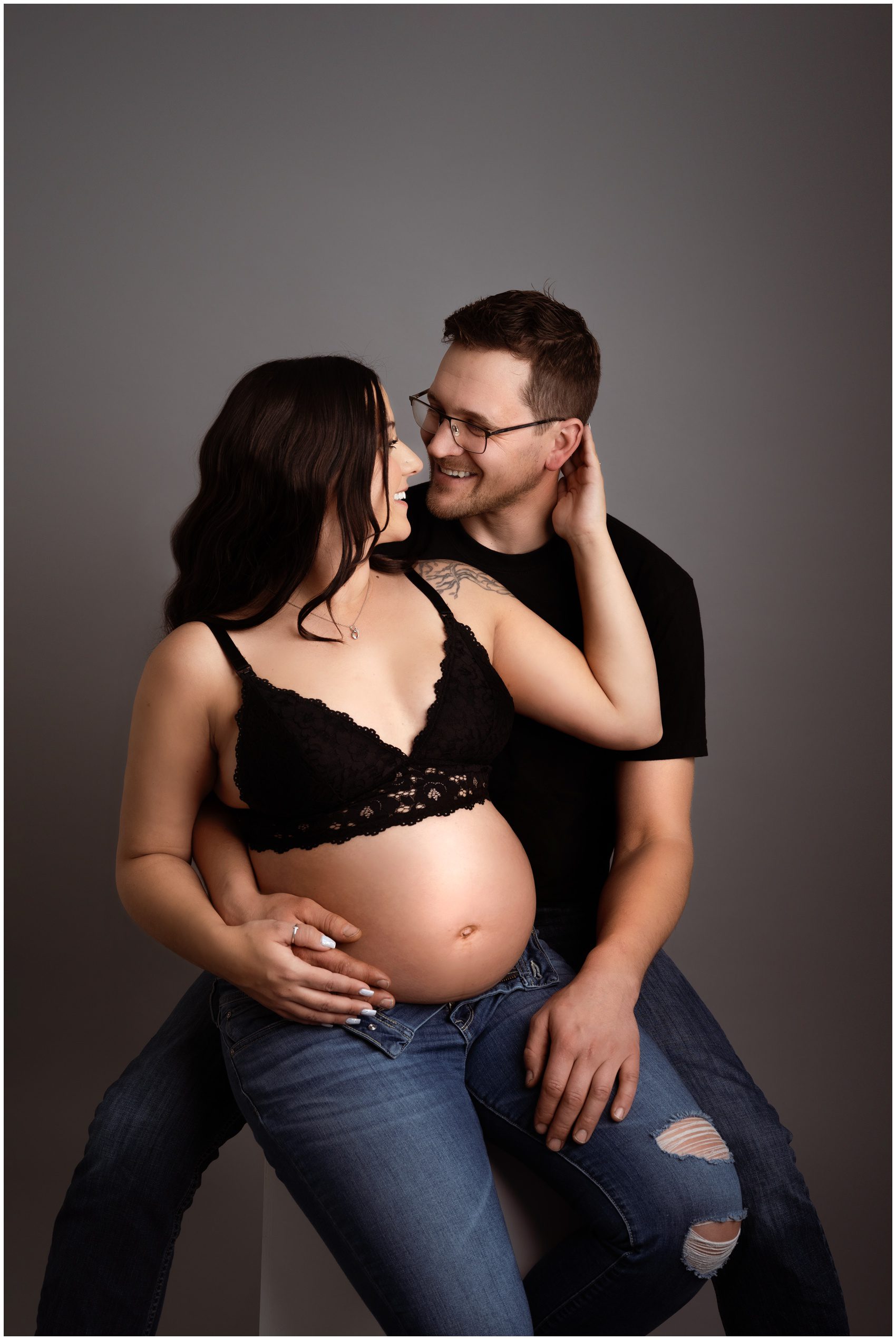 Happy expecting parents sit snuggling on a box in a studio with the bump exposed after meeting edmonton doulas