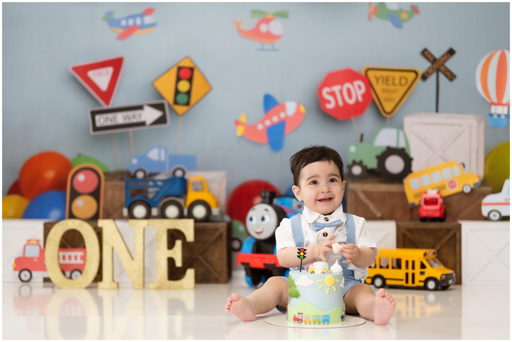 A young toddler in blue suspenders and bowtie sits on a studio floor with a birthday cake