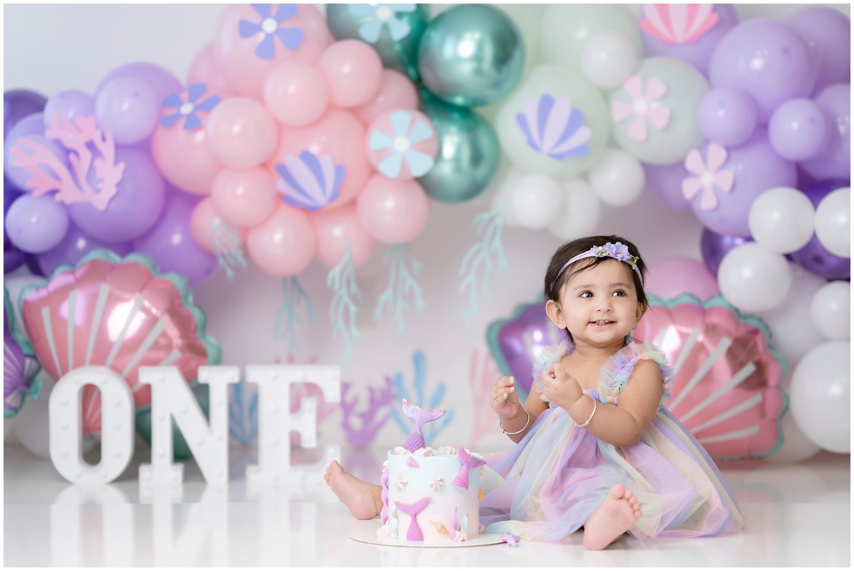 A happy toddler girl ina. colorful dress plays in a mermaid cake in a matching studio