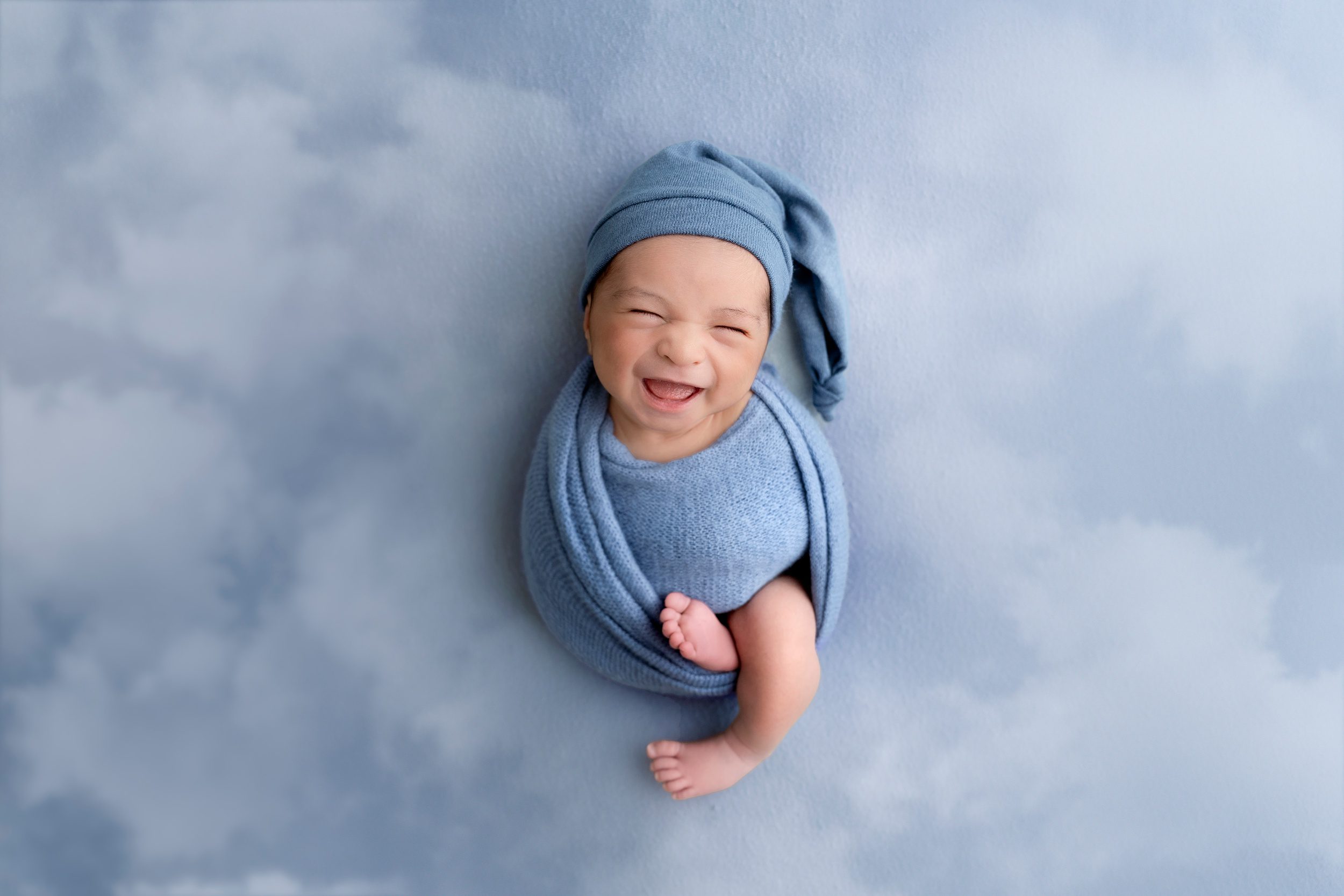 A newborn baby smiles while wrapped in a swaddle with matching night cap on a cloud bed