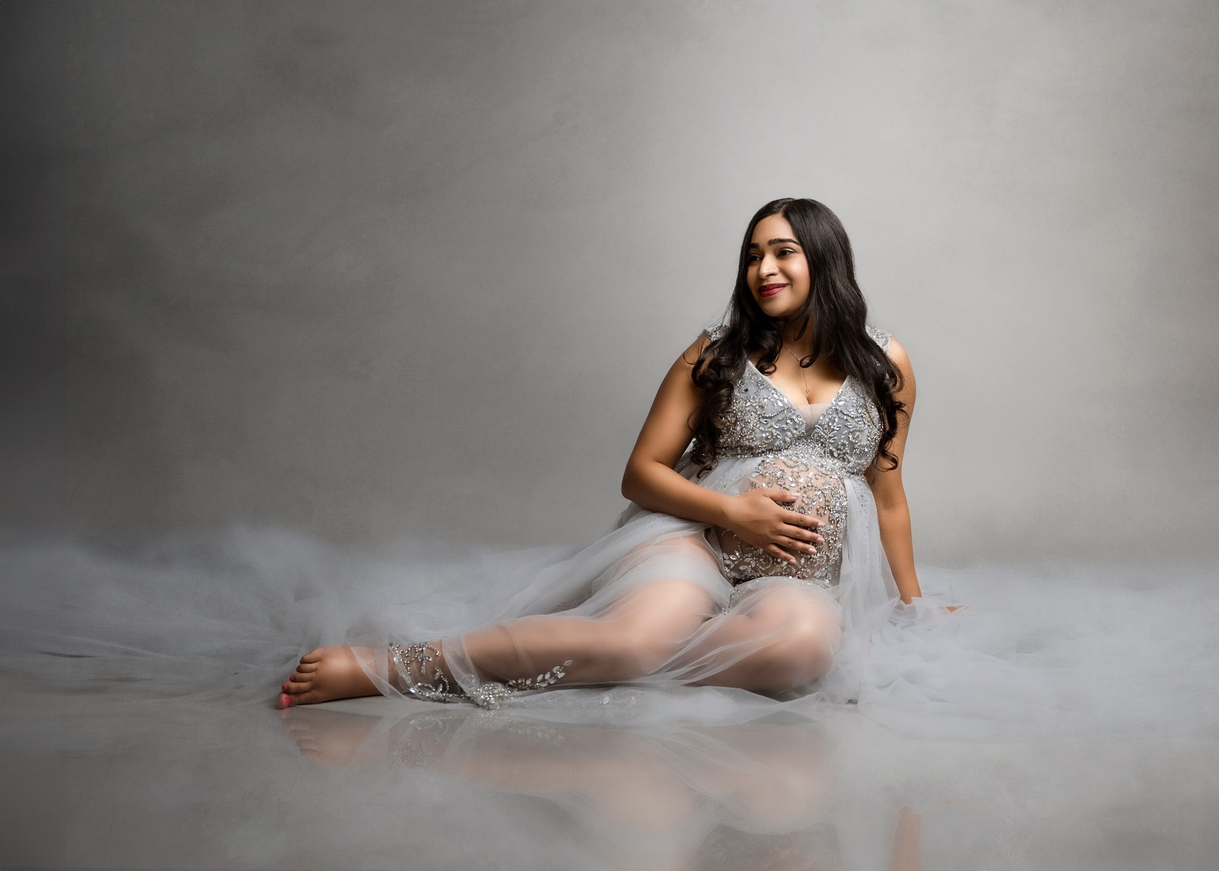 A smiling mother to be sits across the floor of a studio holding her bump and smiling over her shoulder in a silver maternity gown with embroidery after meeting aspen community midwives