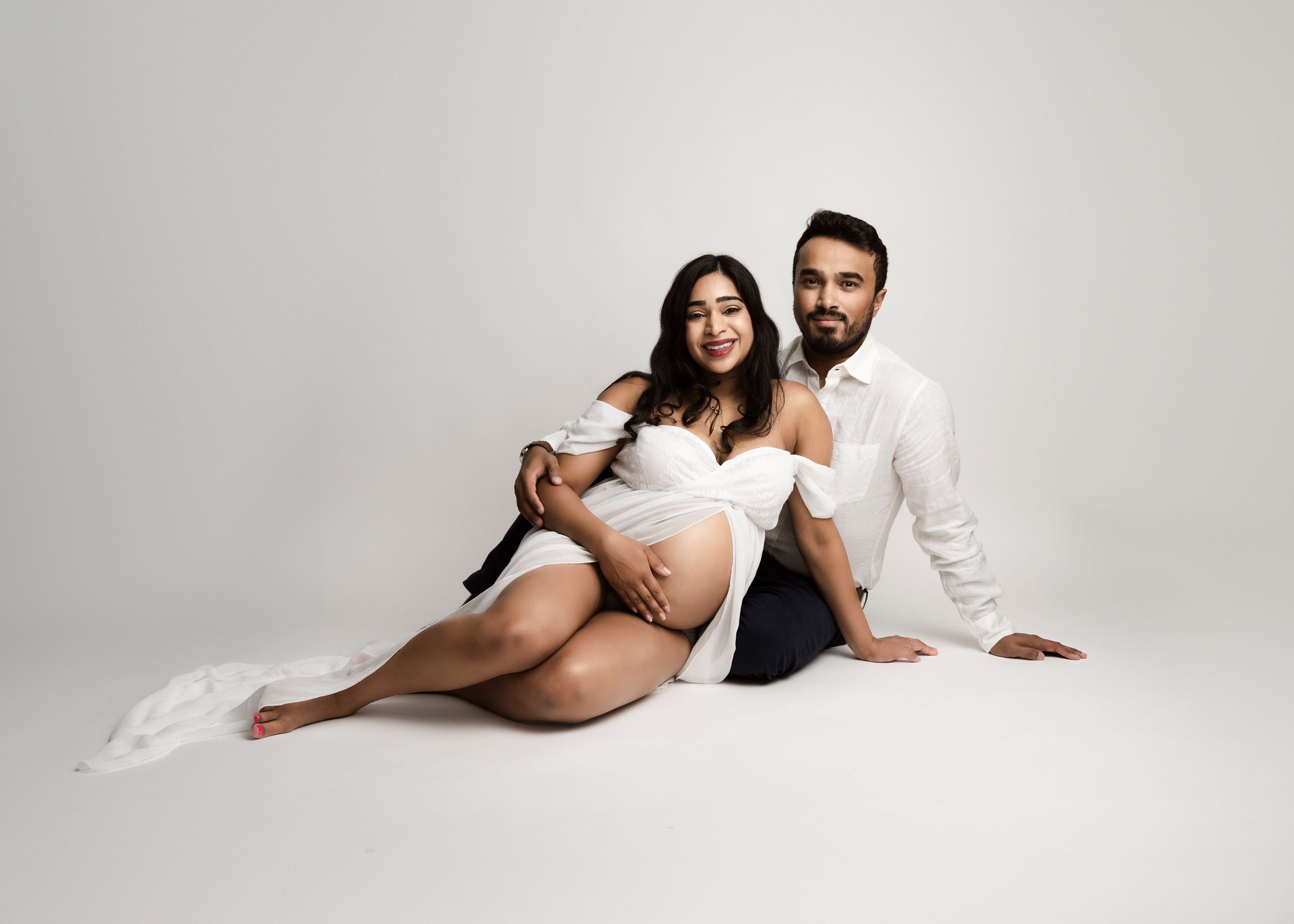 Happy expecting parents sit across the floor of a studio wearing white and smiling after meeting with edmonton area family doulas