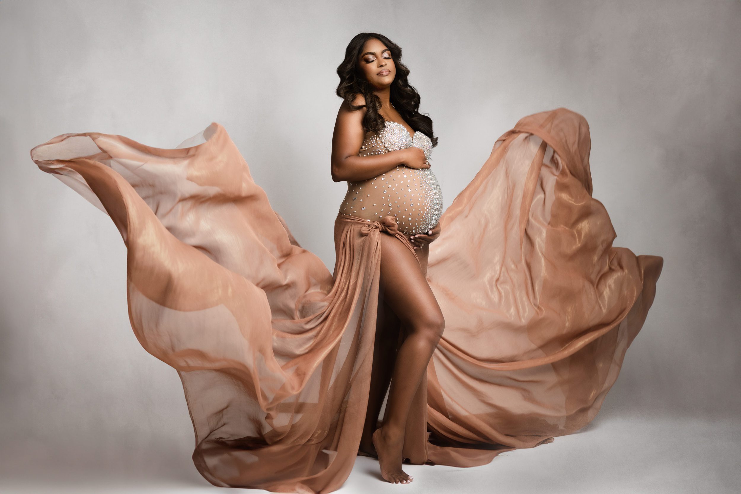 A mom to be stands in a brown and diamond maternity gown as the train flows in the air around her after meeting edmonton midwifery cooperative