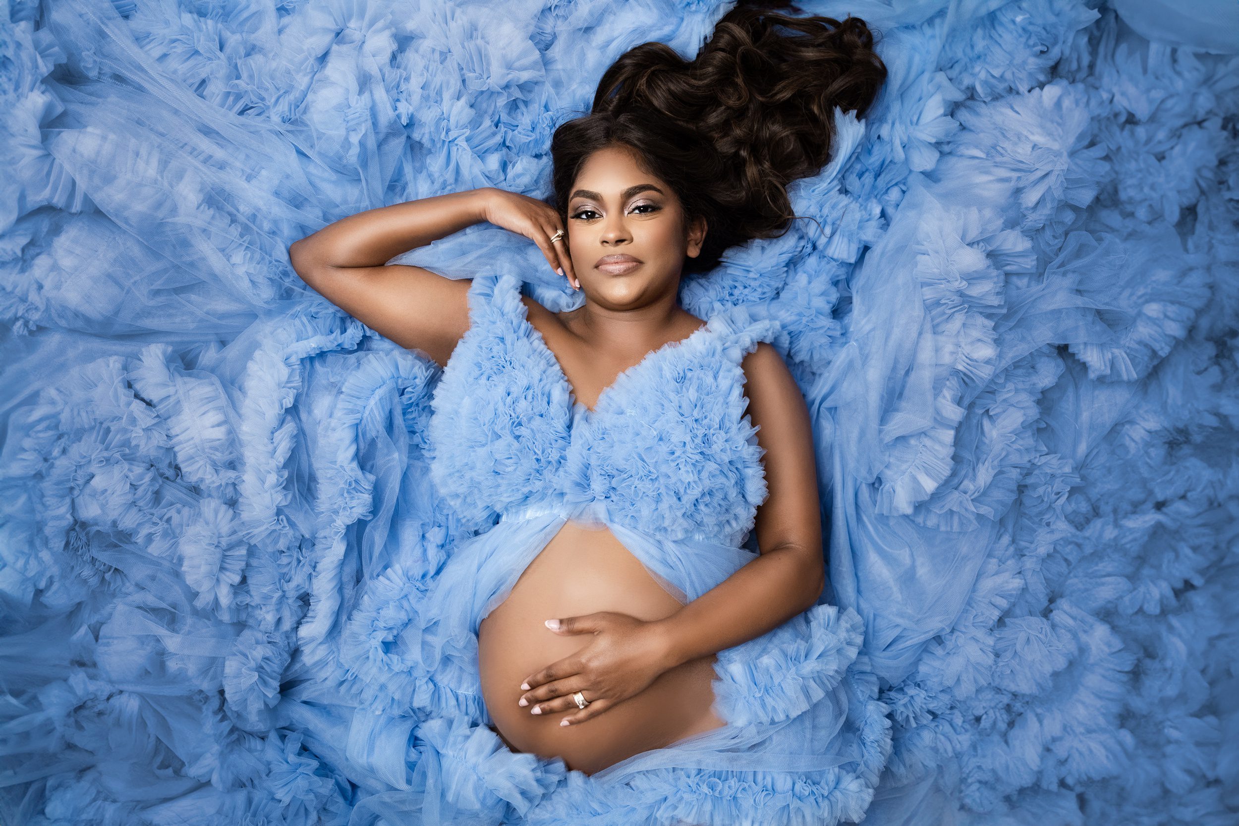 A pregnant woman rests a hand on her bump while laying in a bed of tule in a studio in a matching maternity gown