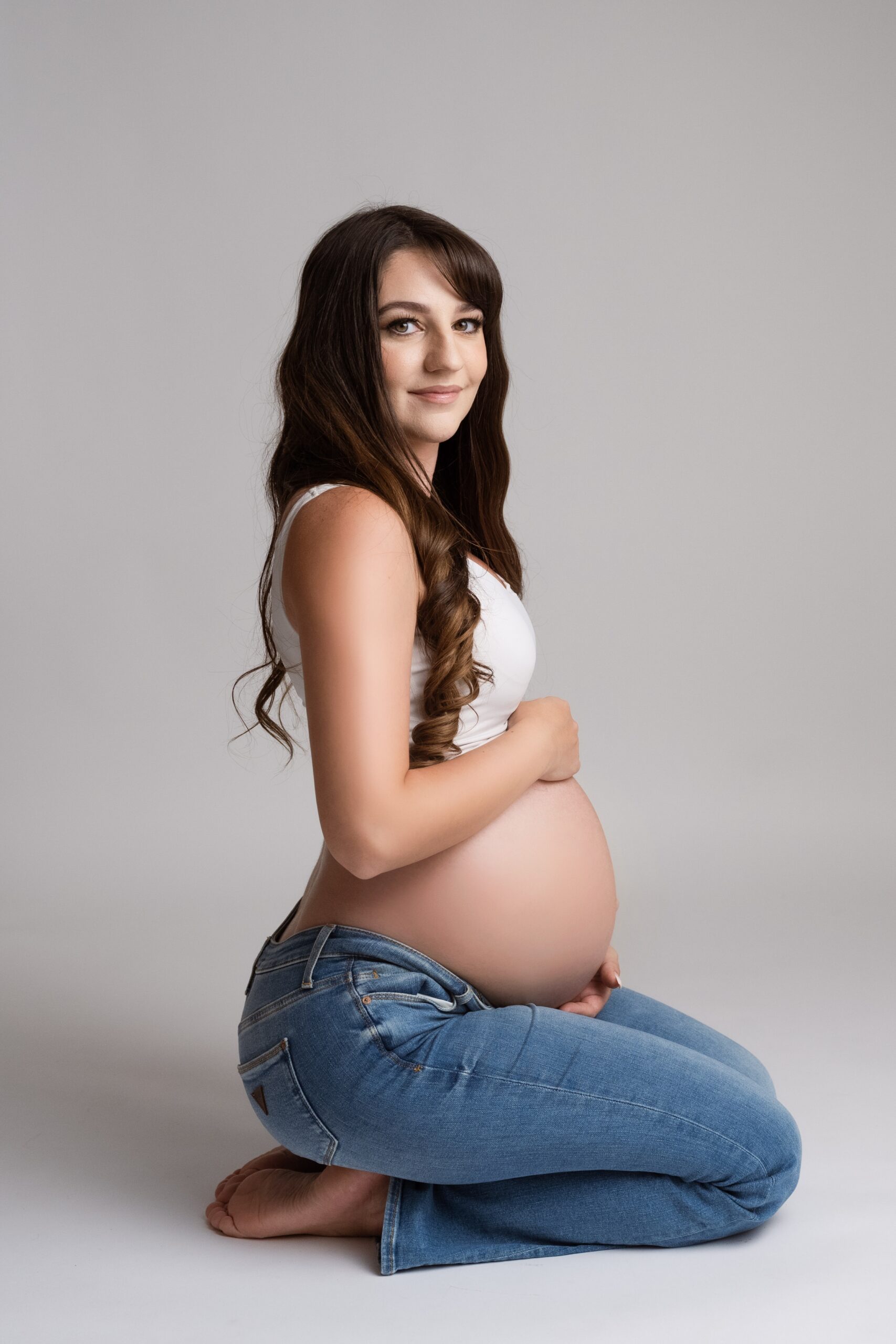 A happy mom to be kneels in a studio wearing jeans and a white top while holding her bump after visiting Sorrel River Ranch