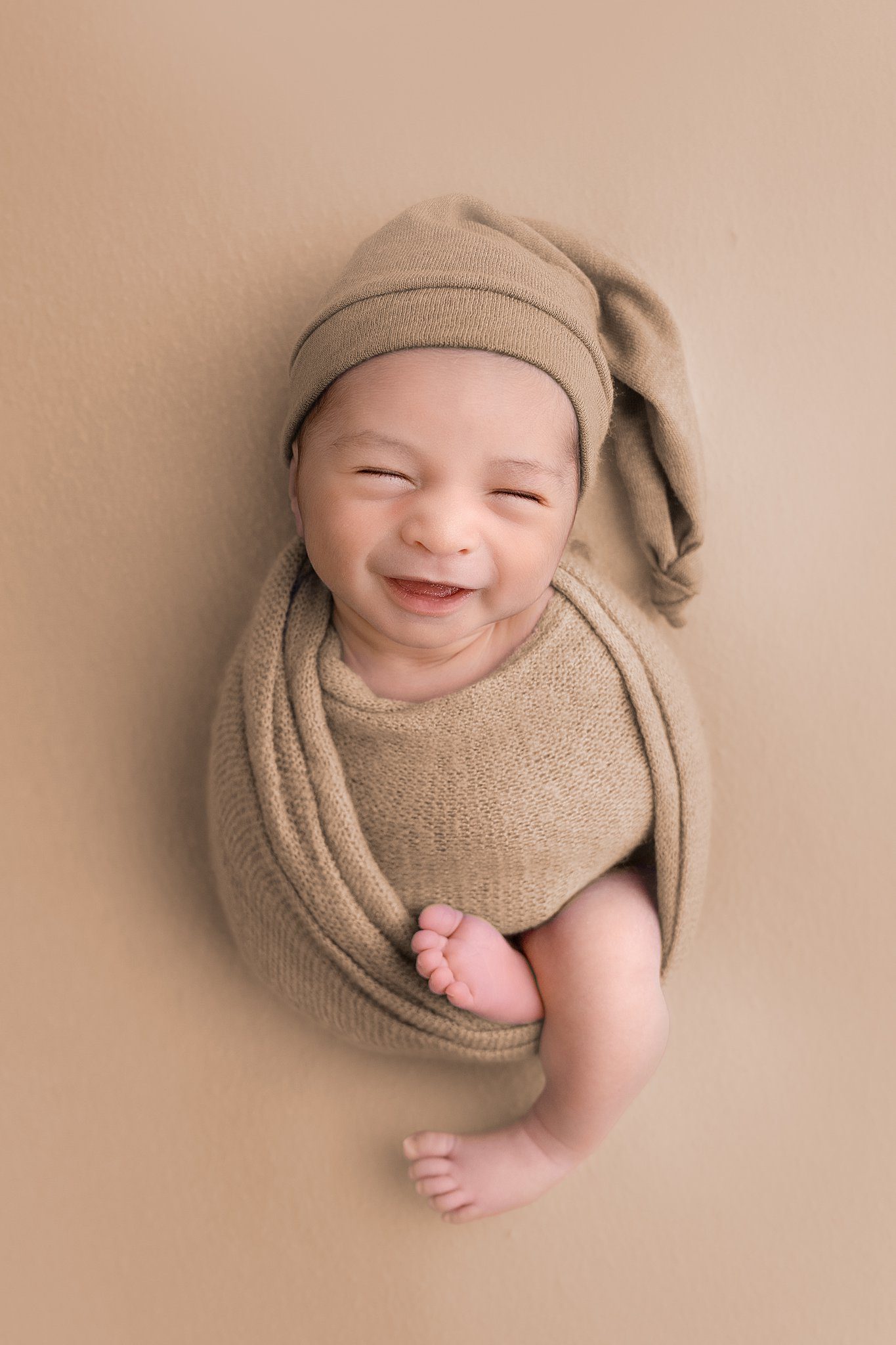 A newborn baby smiles while sleeping in a brown swaddle in a studio